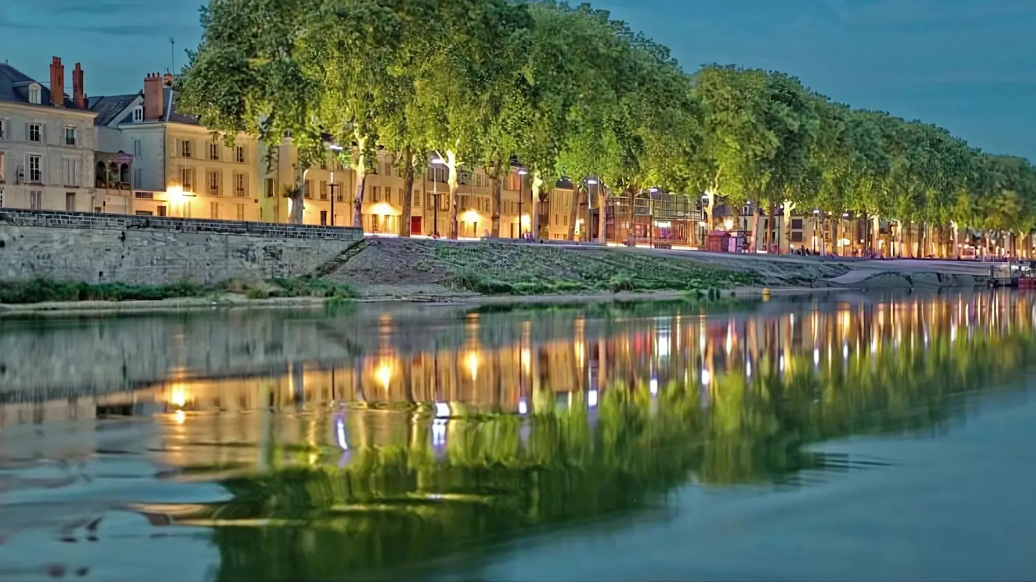 Evening view of a tree-lined riverside with warm lights reflecting on calm water and buildings in the background.