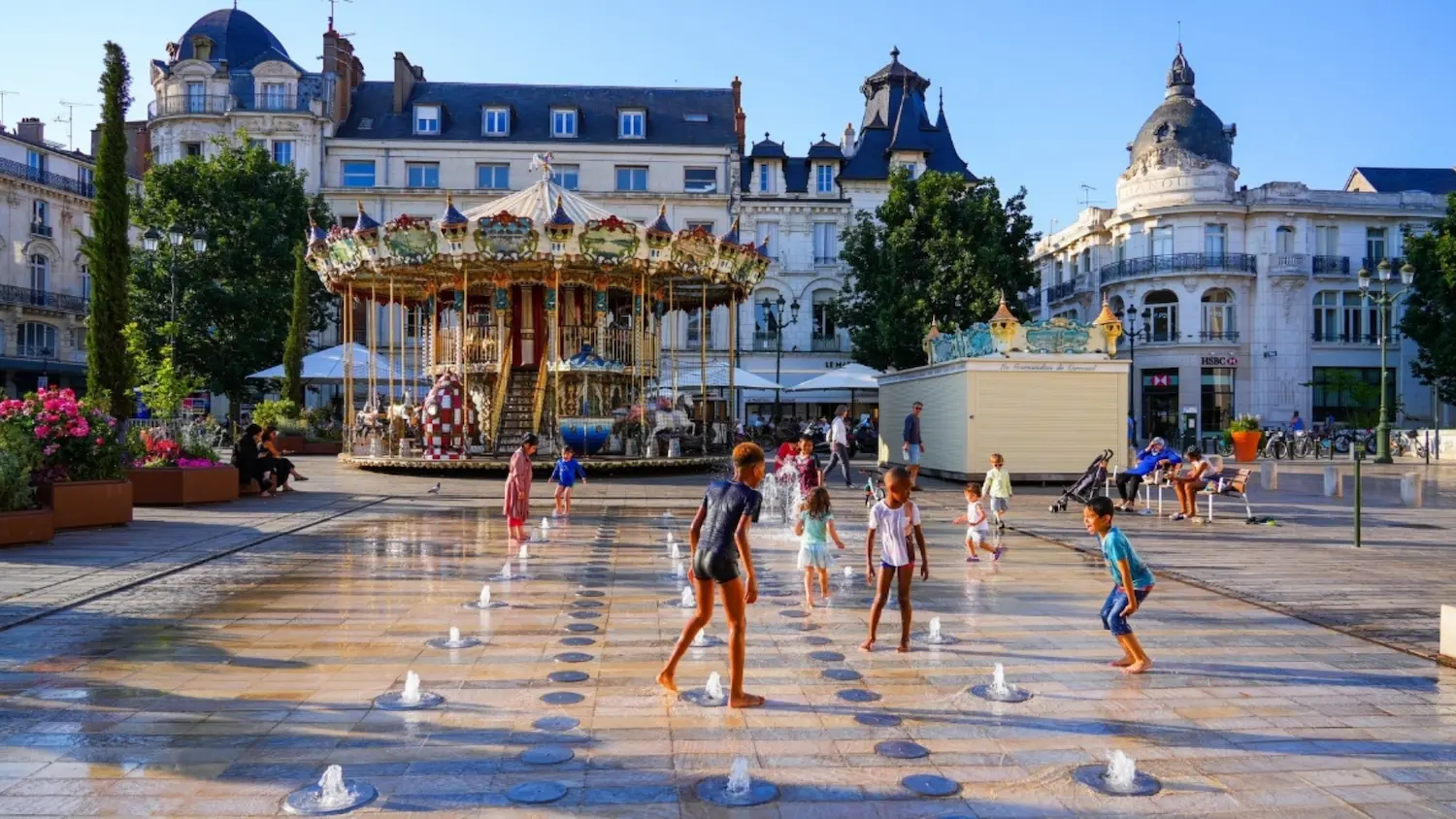 Children playing barefoot in a fountain area with water jets in a European square featuring a vintage carousel and historic buildings.