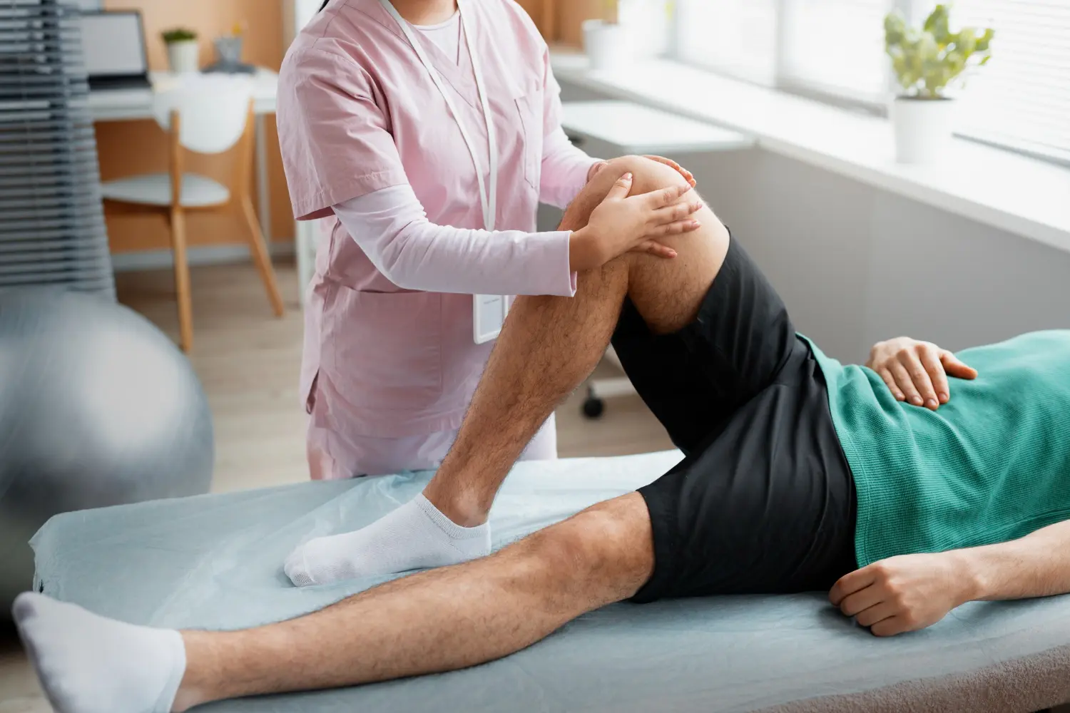 Physical therapist in pink scrubs assisting a man in green shirt and black shorts with knee rehabilitation exercises on a treatment table.