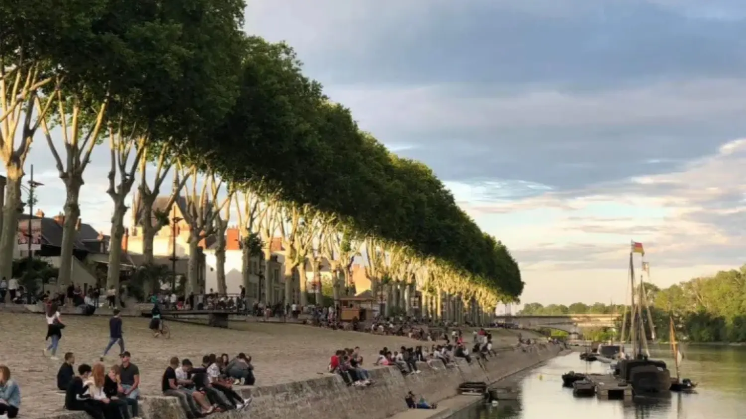 Crowd of people sitting and walking along a riverbank lined with tall trees and boats docked in the water under a partly cloudy sky.