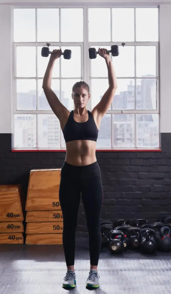 Woman in black workout attire lifting dumbbells overhead in a gym with large windows and stacked wooden boxes in the background.