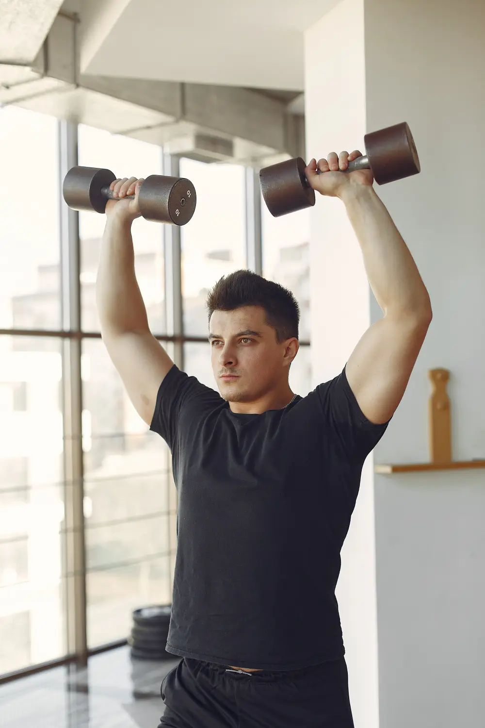 Man in black shirt lifting two 10-pound dumbbells overhead in a bright gym with large windows.