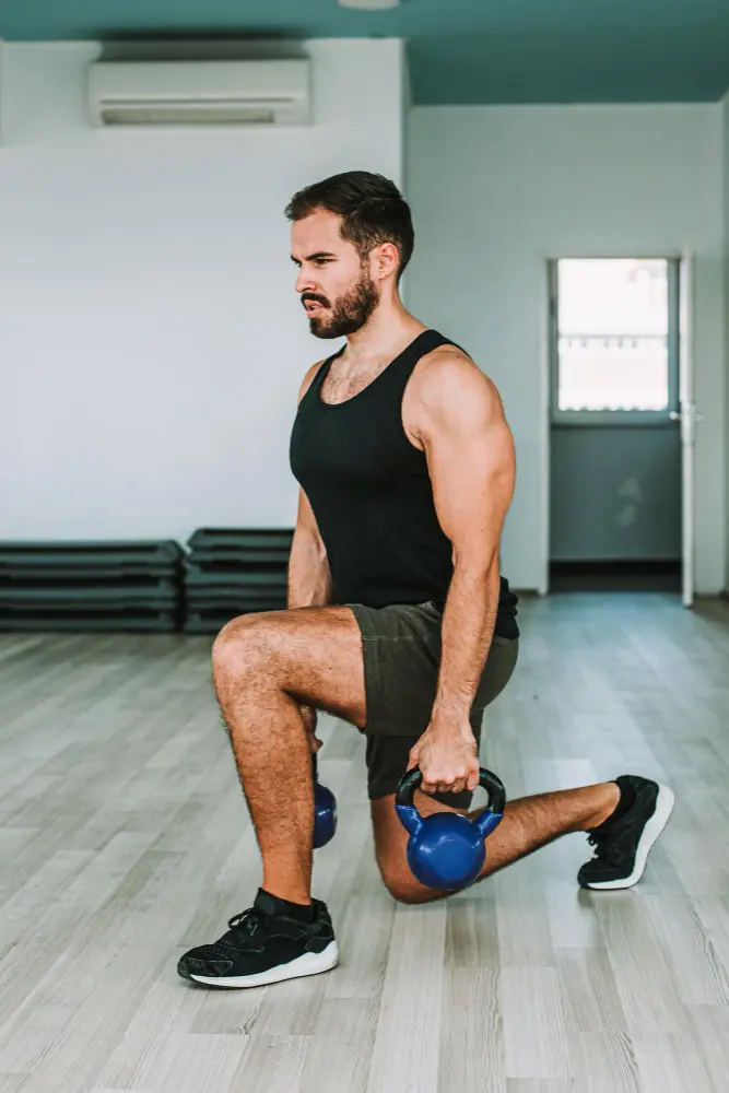 Man performing lunges indoors while holding two blue kettlebells.