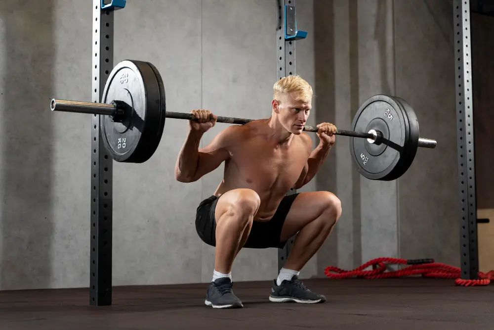 Shirtless man performing a deep barbell squat in a gym squat rack.