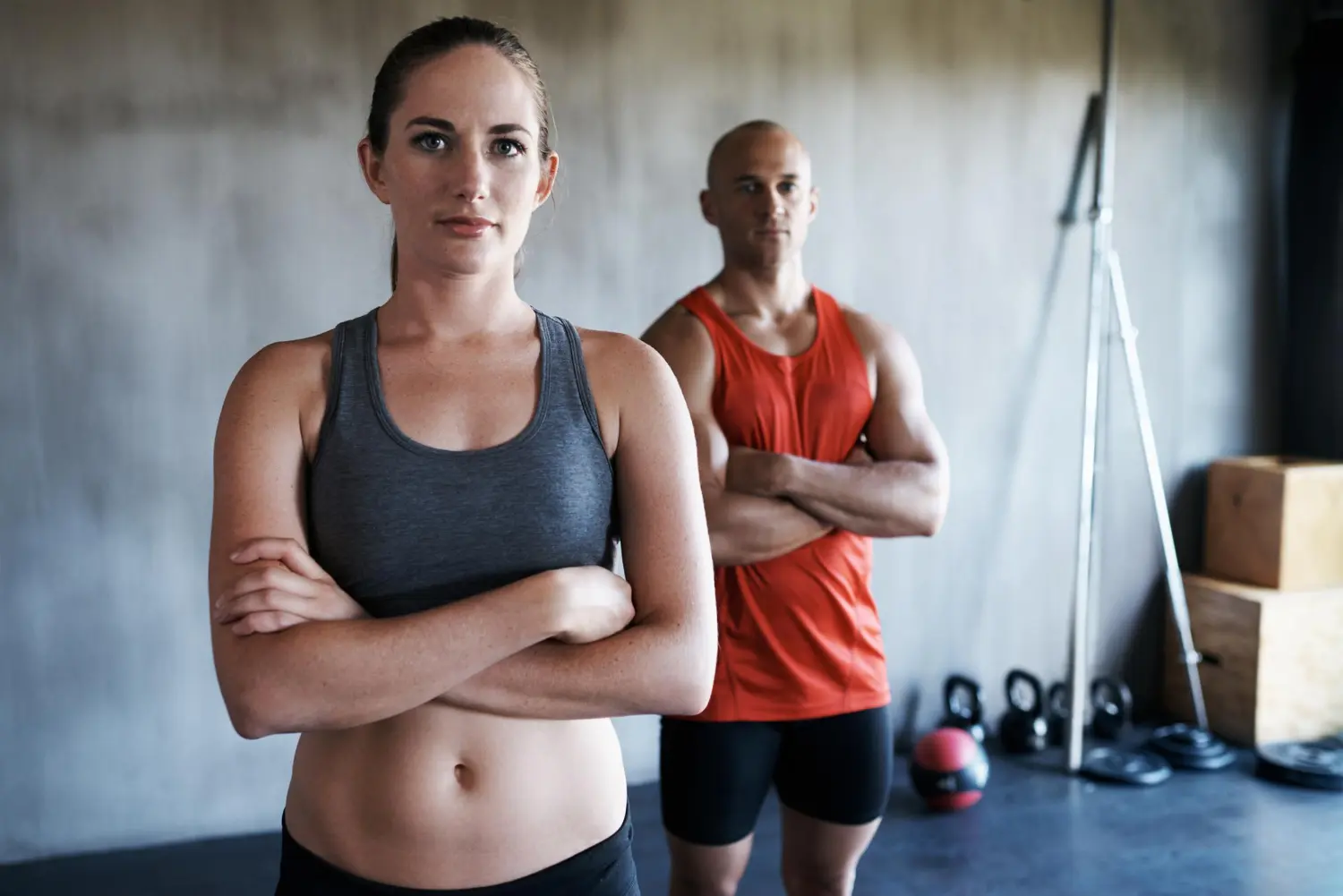 Fit woman in gray sports bra and man in red tank top standing with arms crossed in a gym.