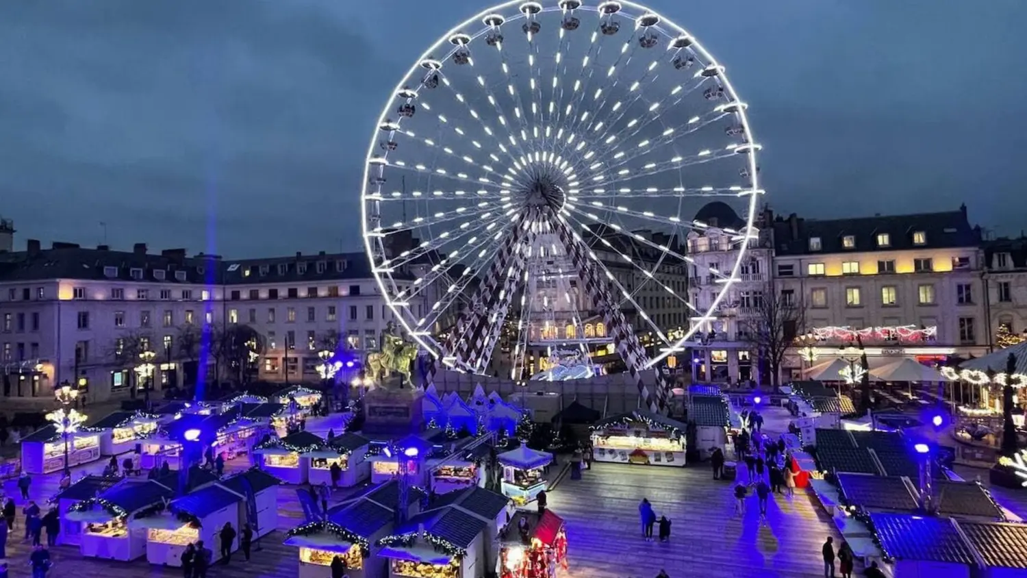 Night scene of a lively outdoor market with illuminated stalls and a large, brightly lit Ferris wheel in the background.