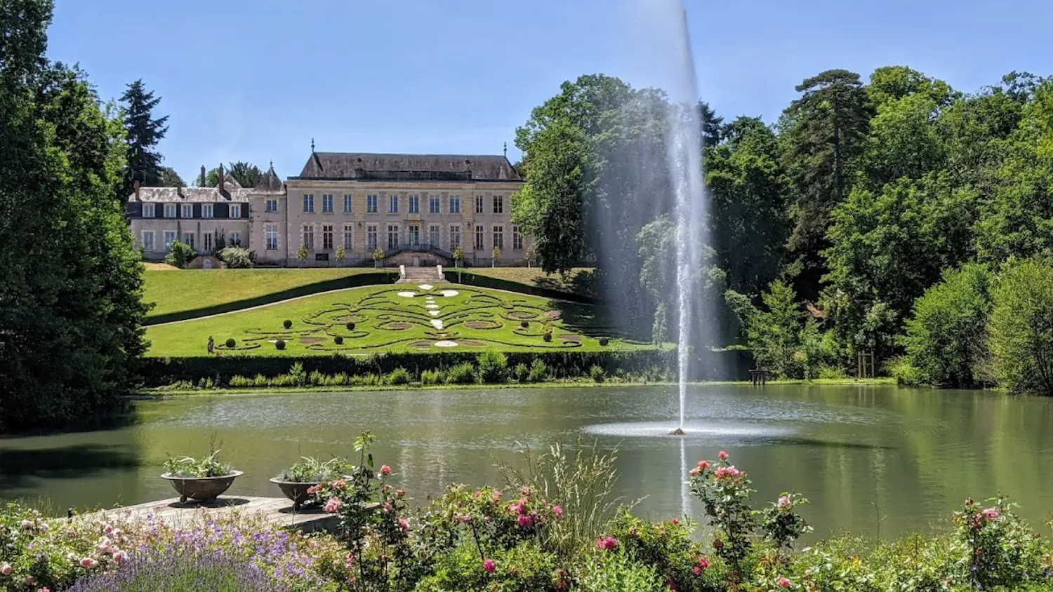 Historic mansion with formal gardens, a pond with a tall water fountain, and blooming flowers in the foreground under a clear blue sky.