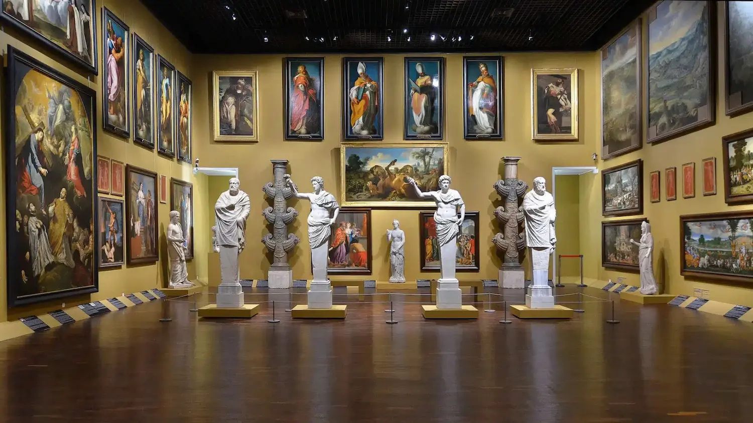Museum gallery room with marble statues on pedestals and various framed classical paintings on yellow walls.