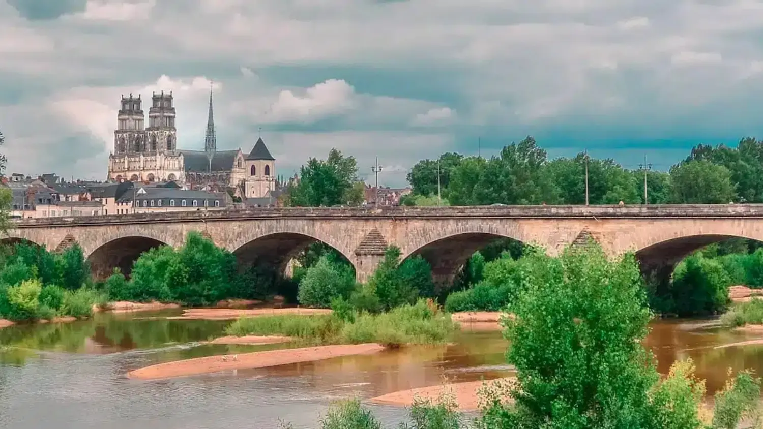 Stone arch bridge over a river with lush green trees and historic cathedral buildings in the background under a cloudy sky.
