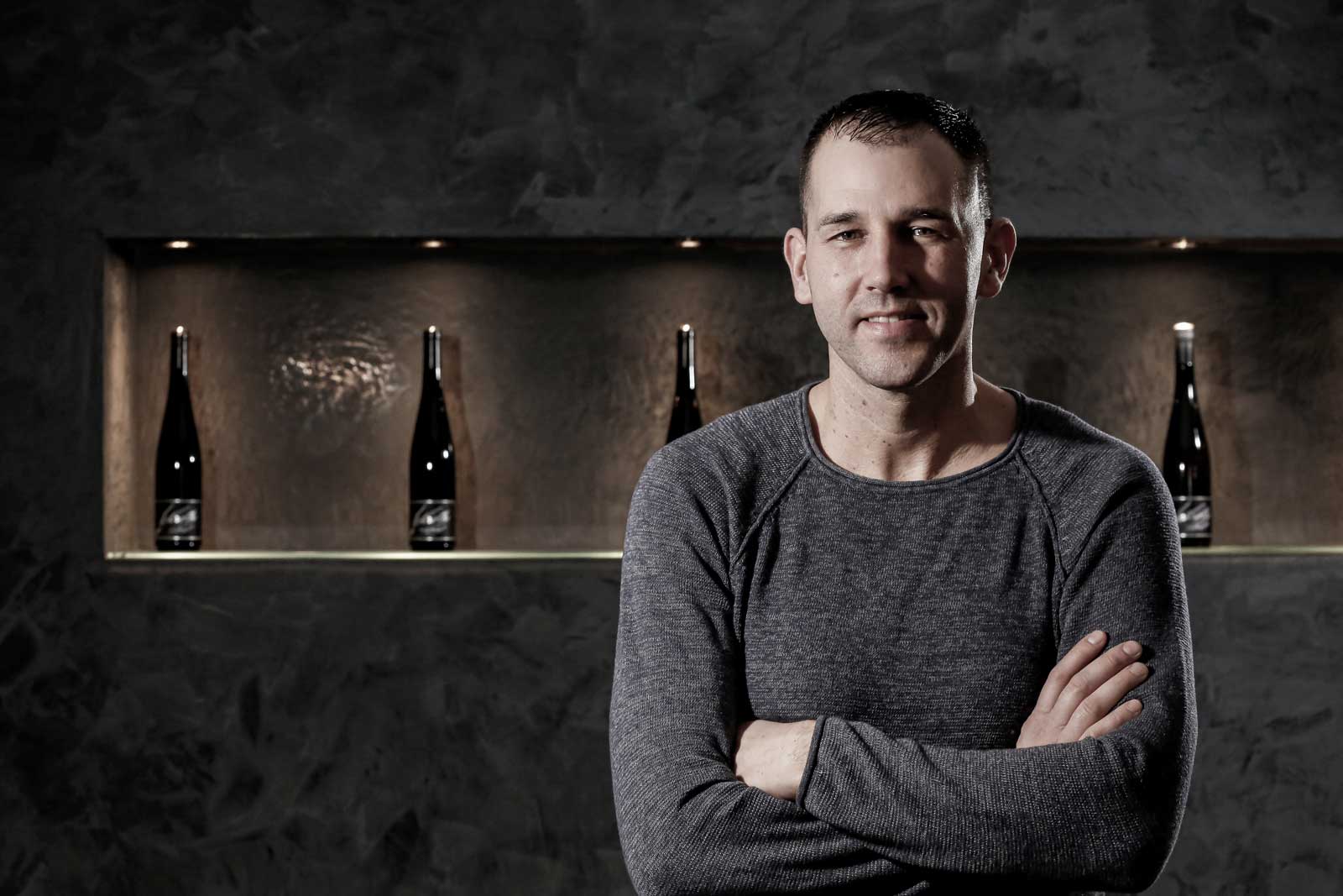 Michael Schroth vor dunklem Hintergrund mit vier Weinflaschen auf einem Regal.