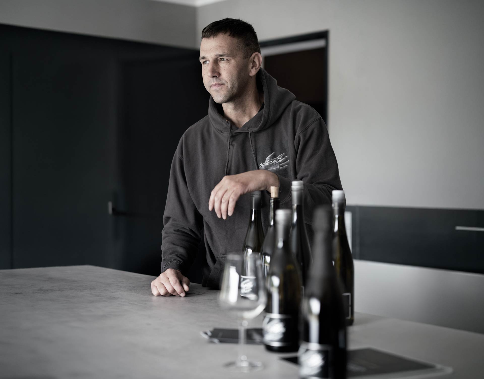 Michael Schroth in Kapuzenpullover lehnt an Tisch mit mehreren Weinflaschen und einem Weinglas in der Vinothek des Weinguts Schroth in Asselheim Pfalz.