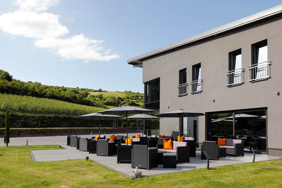 Weingut Schroth in Asselheim mit moderner Terrasse mit Sitzmöbeln und Sonnenschirmen neben einem grauen Gebäude vor Weinbergen unter blauem Himmel.