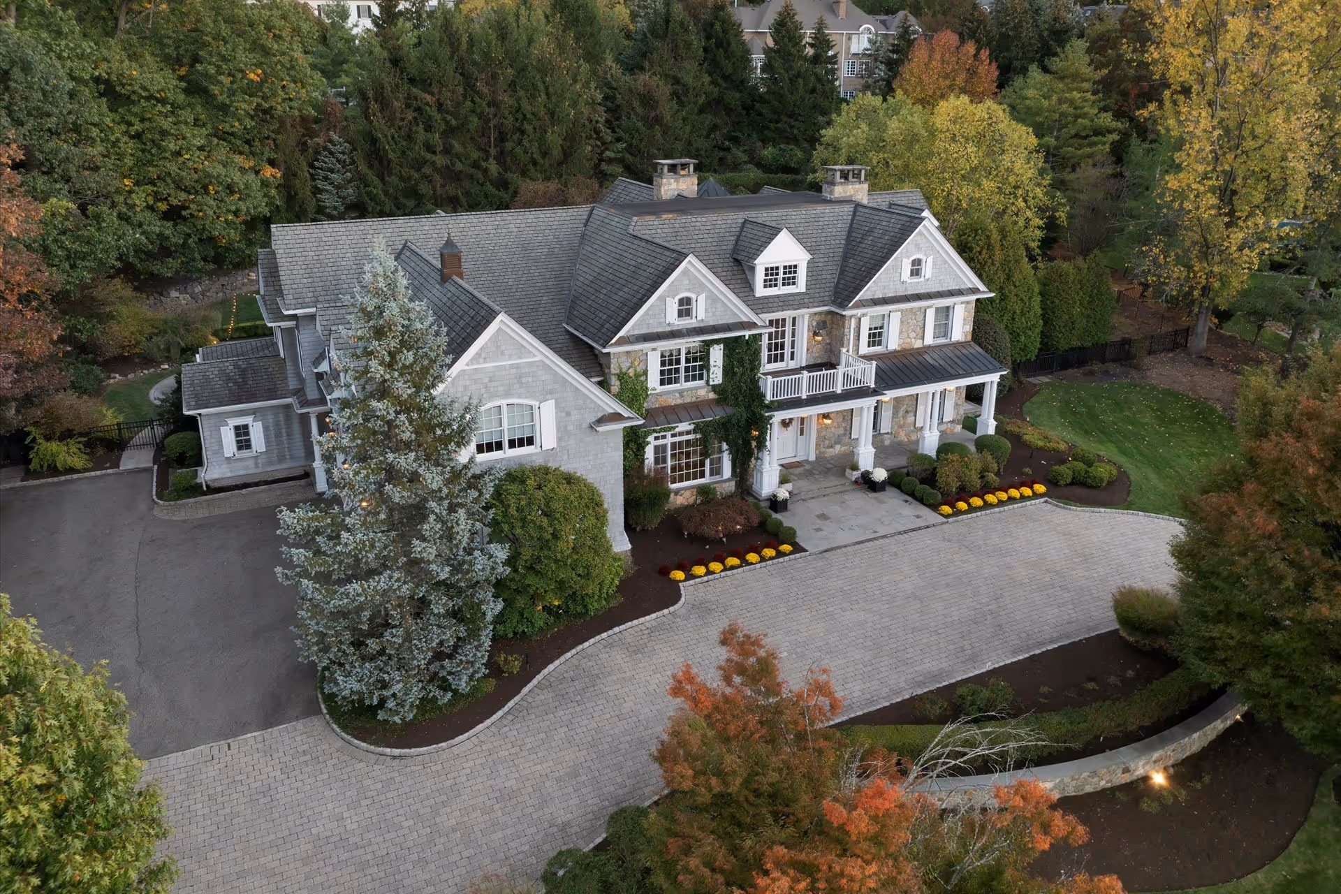 A large, elegant two-story house with a gray roof and stone exterior surrounded by neatly landscaped greenery and trees in autumn colors.