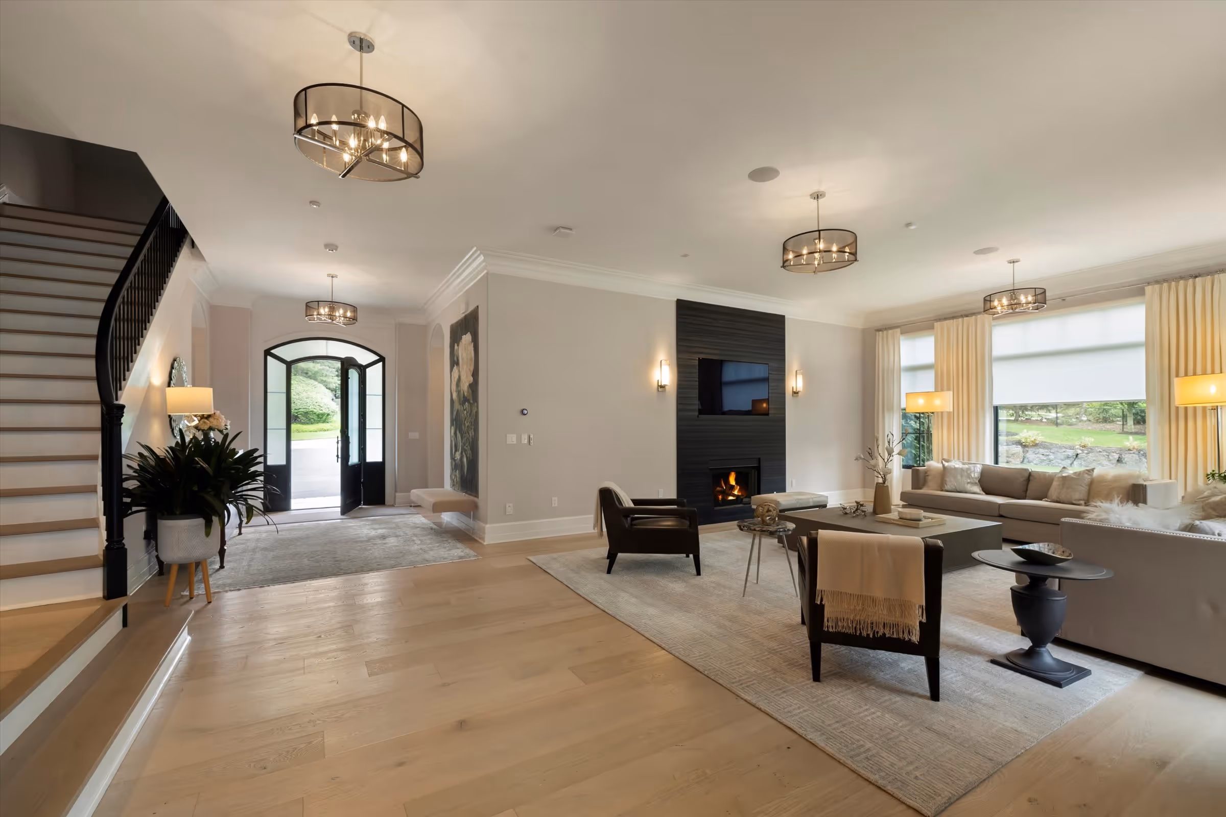 Spacious modern living room with light hardwood floors, neutral sofas, black armchairs, fireplace, and large windows with curtains.