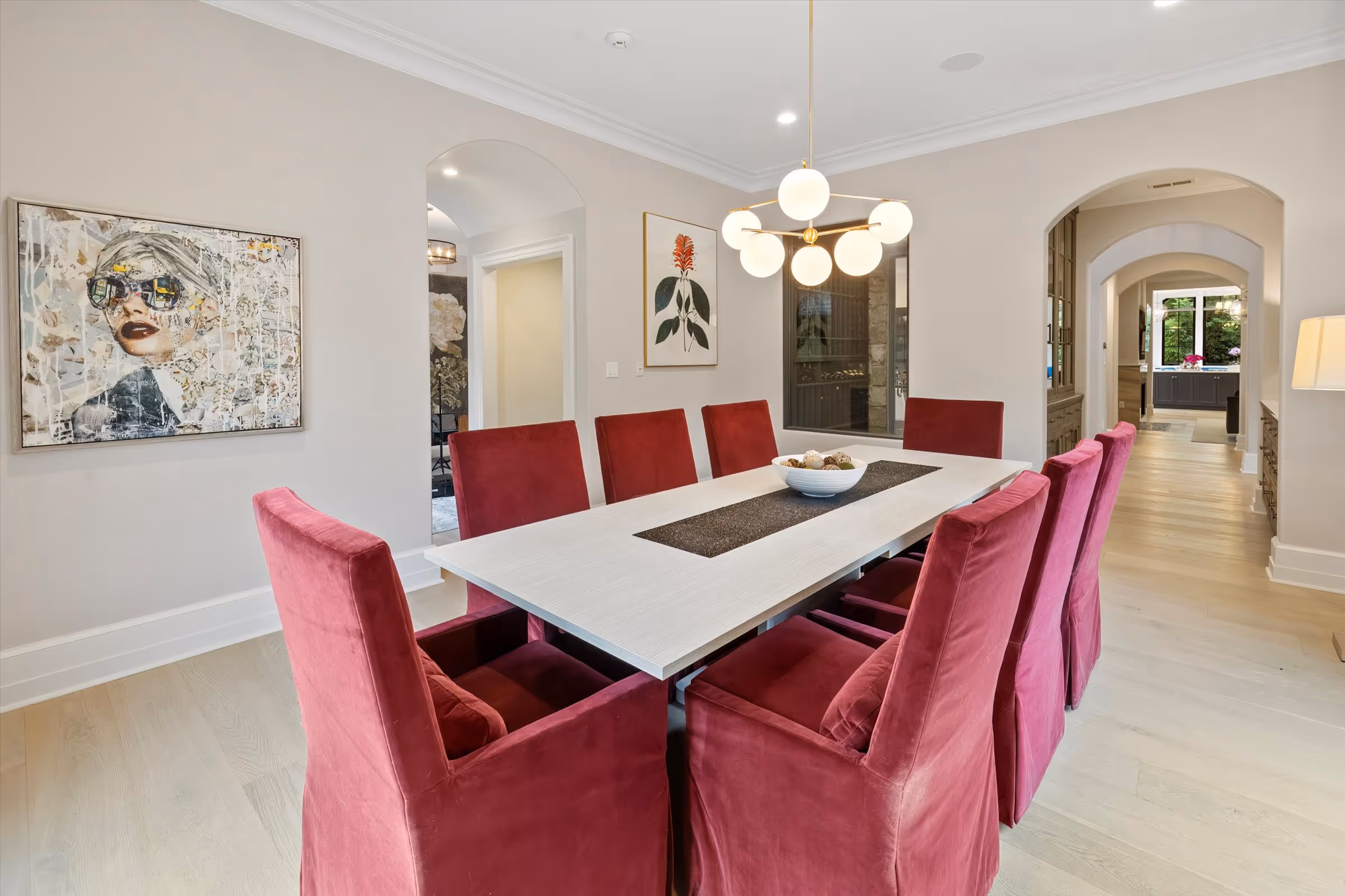 Modern dining room with a white rectangular table surrounded by eight red upholstered chairs and spherical chandelier above.