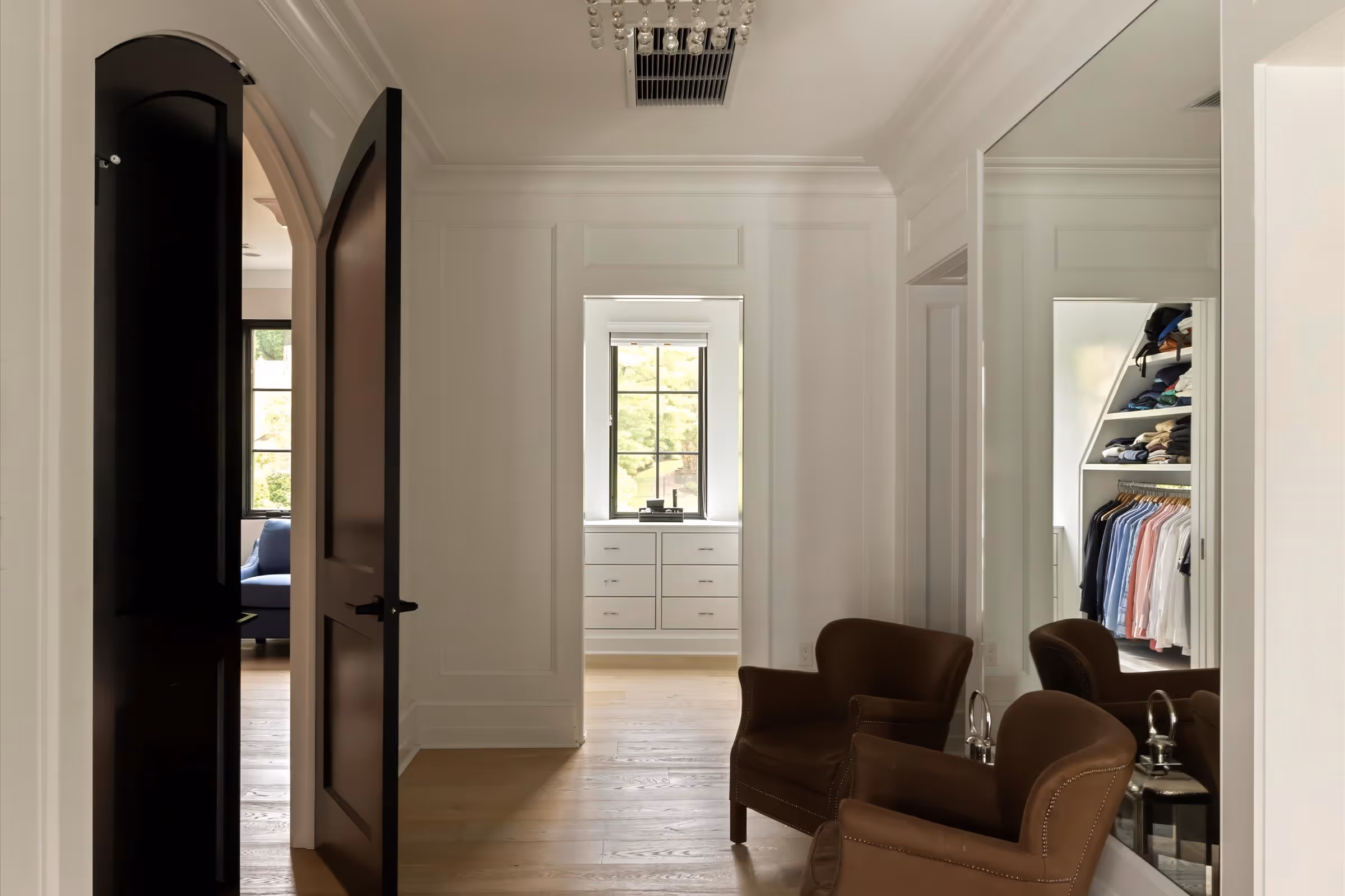Interior hallway with open dark wooden door, two brown armchairs, large mirror, and a window with white drawers beneath it.