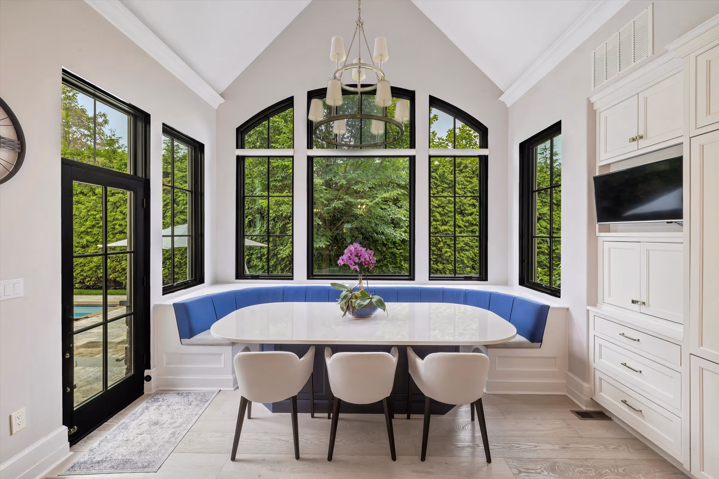 Modern dining area with a white table, three white chairs, a blue cushioned bench along large black-framed windows overlooking greenery, and a hanging chandelier.