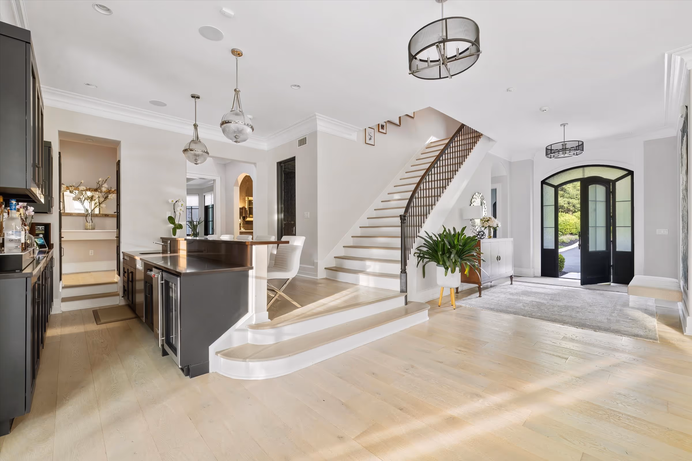 Bright open foyer with light wood flooring, a staircase with black railing, dark kitchen island, modern pendant lights, and black front doors.