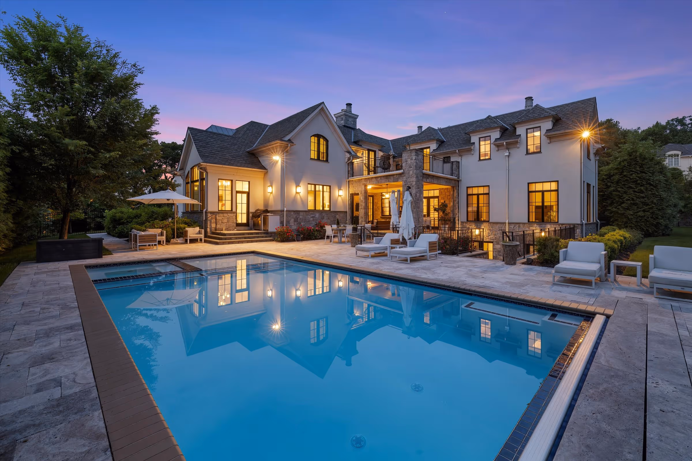 Elegant two-story house with lit windows and a swimming pool reflecting the building at dusk.