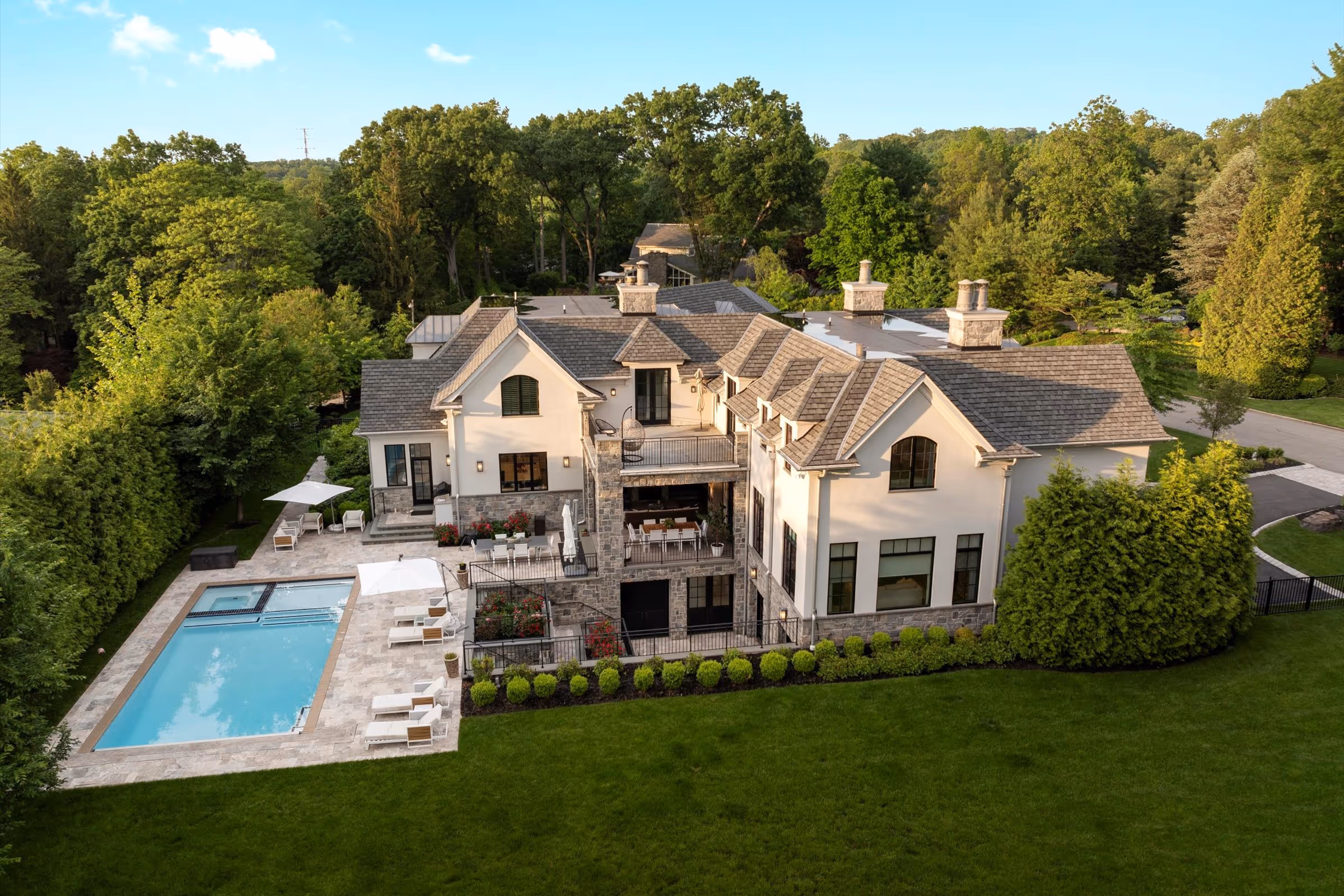 Large modern two-story house with stone and white exterior, featuring multiple balconies, a swimming pool, and a spacious patio surrounded by trees and greenery.