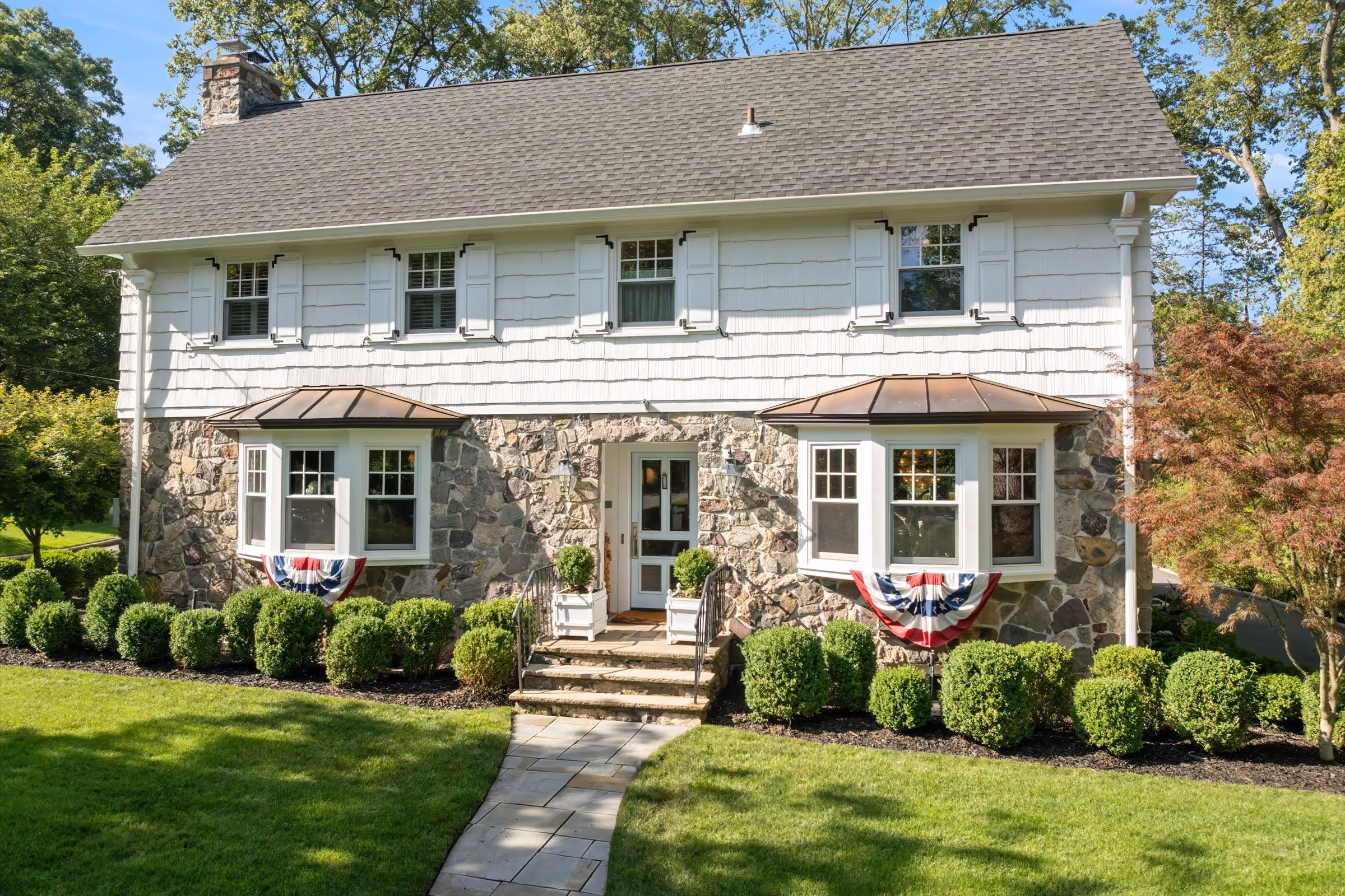 Two-story house with stone facade on the ground floor, white siding above, symmetrical bay windows with flags, and neatly trimmed bushes in front.