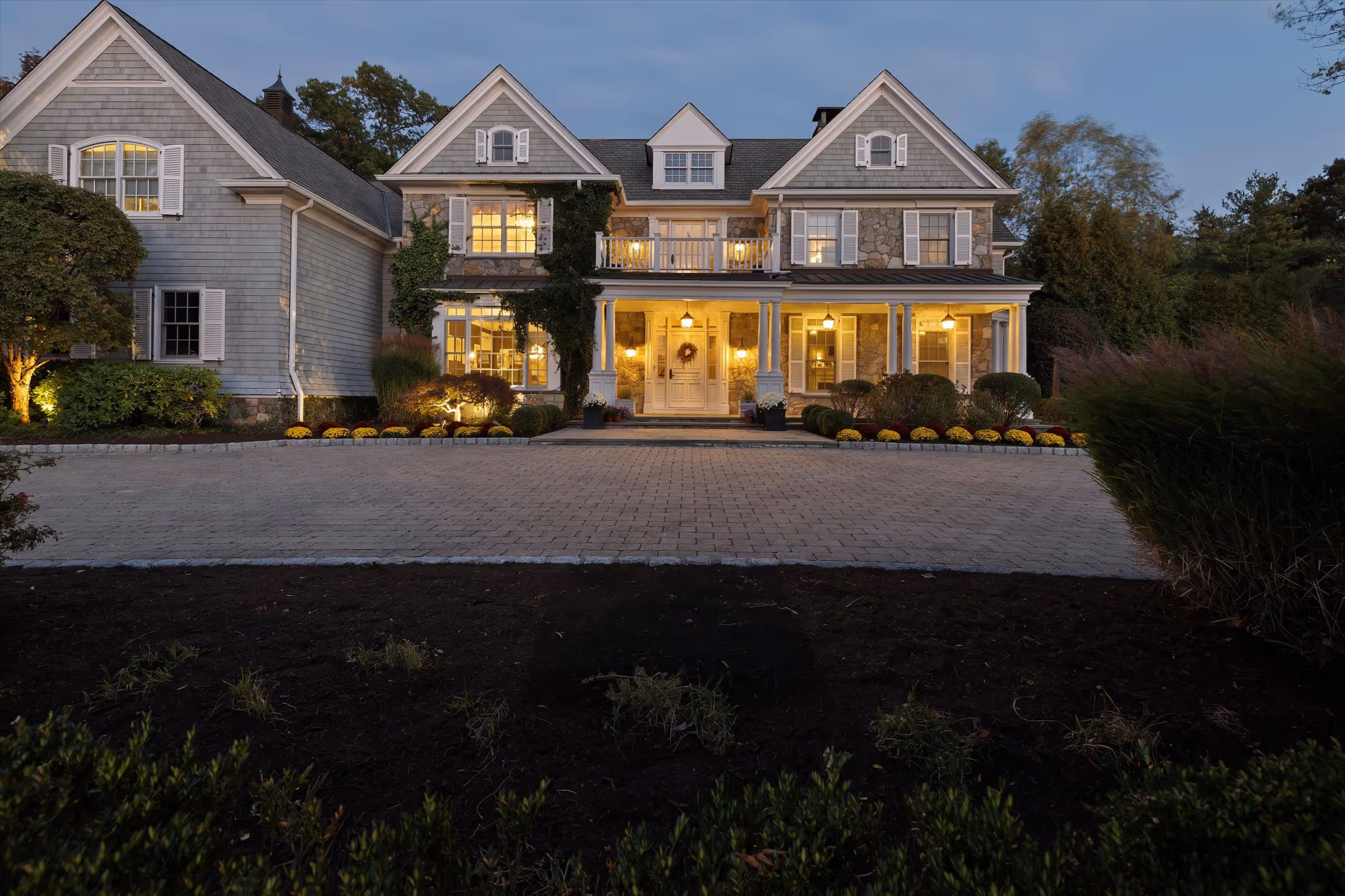 Large two-story house with stone and gray siding exterior, lit warmly with porch lights during twilight.
