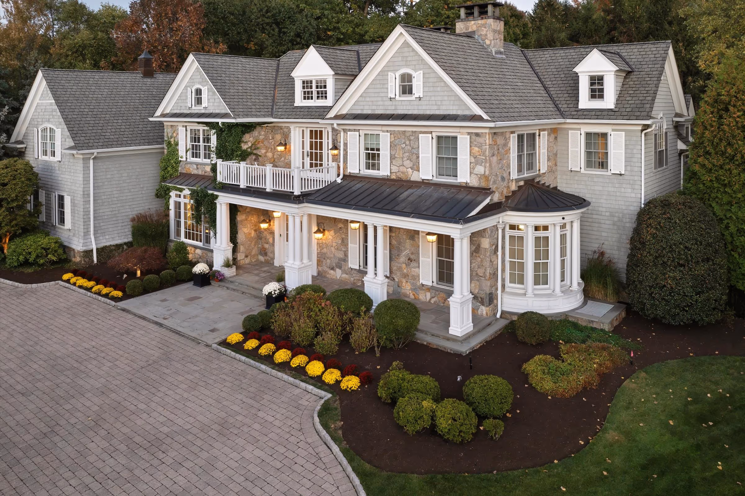 Large two-story house with stone and gray siding exterior, multiple gabled roofs, white window shutters, front porch with columns, and landscaped garden with flowers and shrubs.