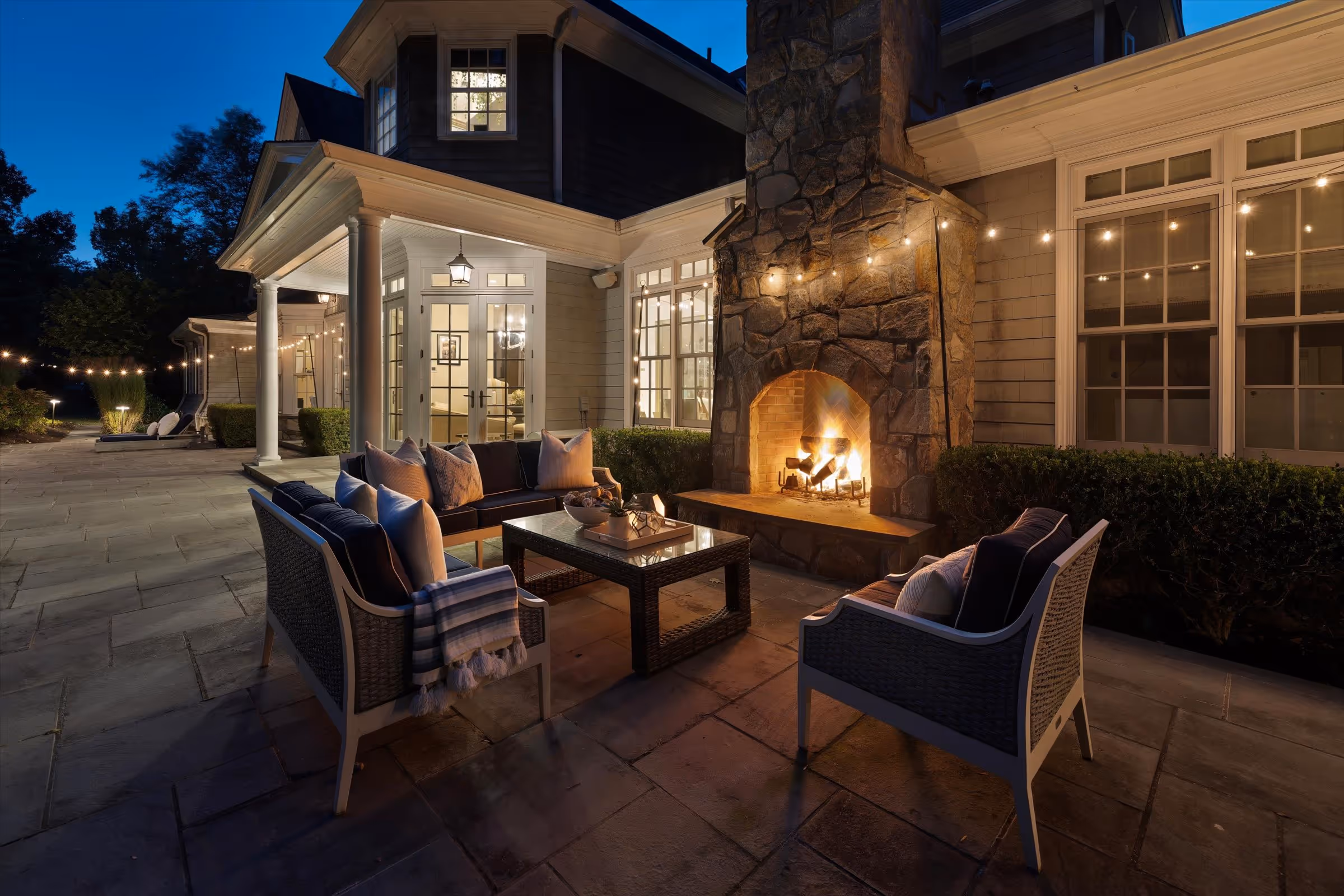 Cozy outdoor patio at night with cushioned seating around a stone fireplace and string lights overhead.