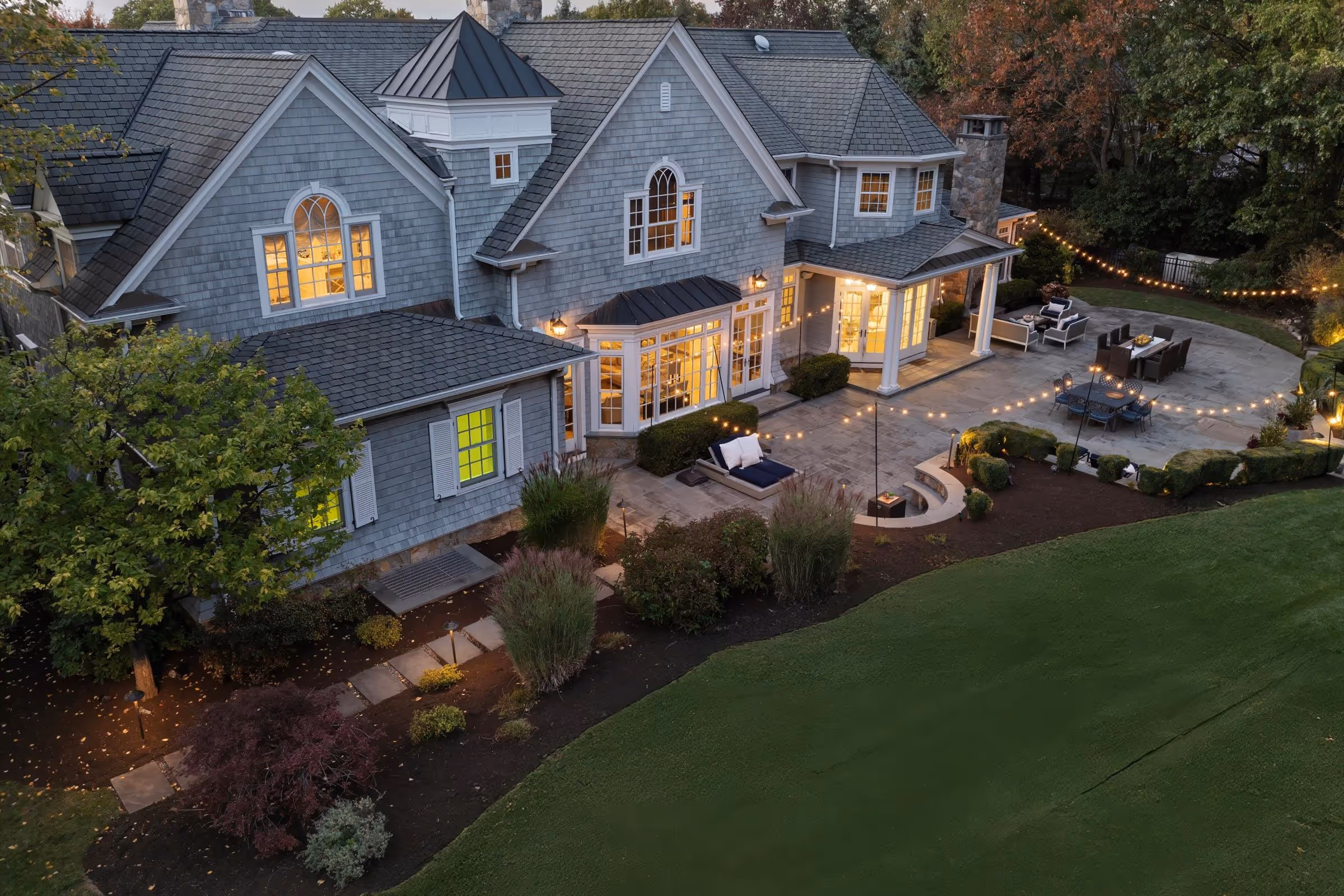 Large gray house with lit windows, a spacious stone patio with outdoor seating, string lights, and surrounding trees and lawn at dusk.