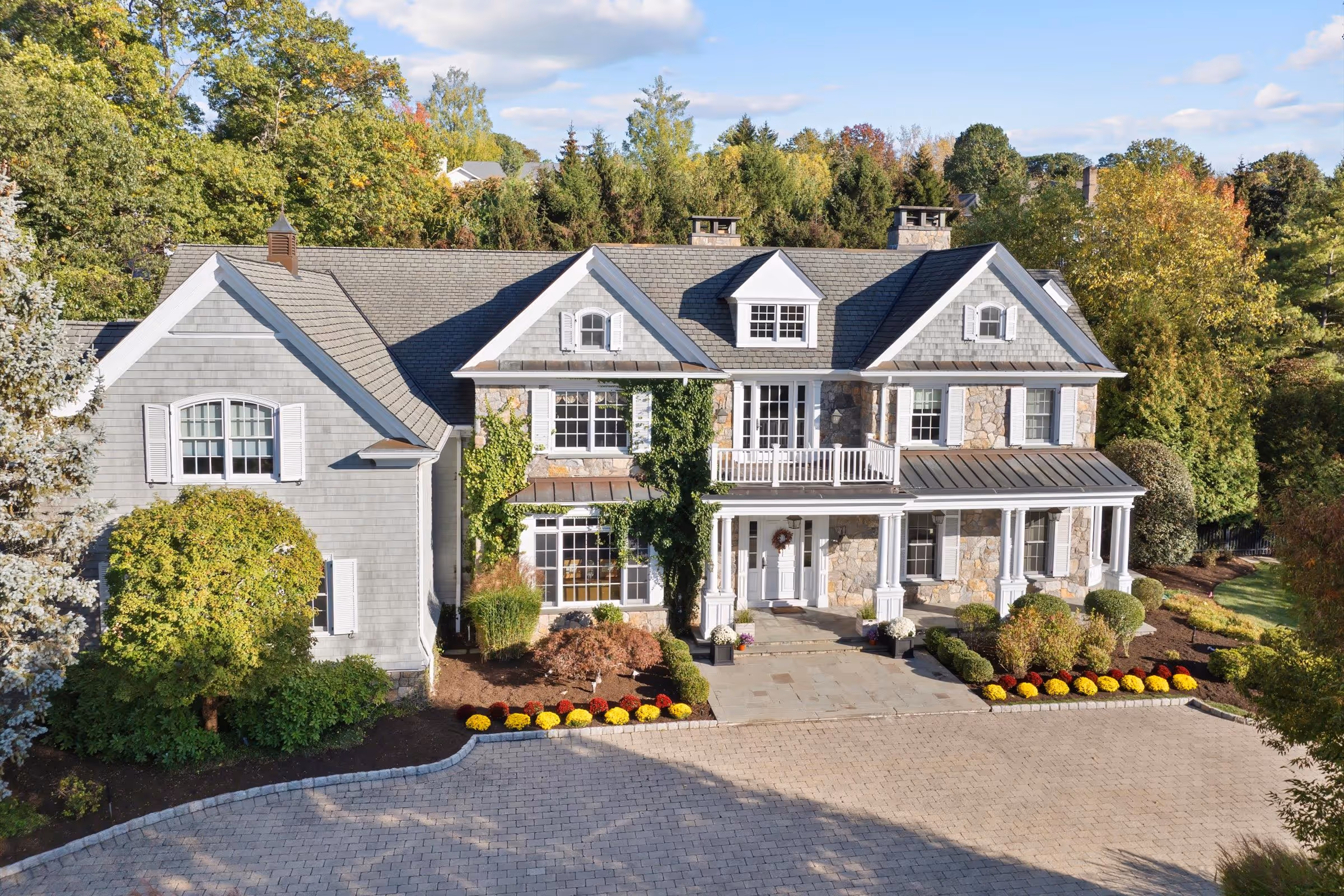 Large two-story house with gray stone and siding exterior, white shutters, a spacious front porch, and landscaped garden with colorful flowers.