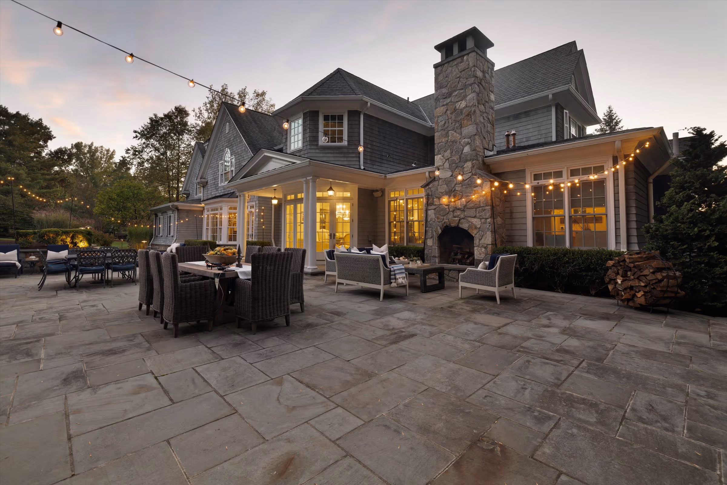 Outdoor patio of a large house at dusk with string lights, stone fireplace, dining table with chairs, and cushioned seating.