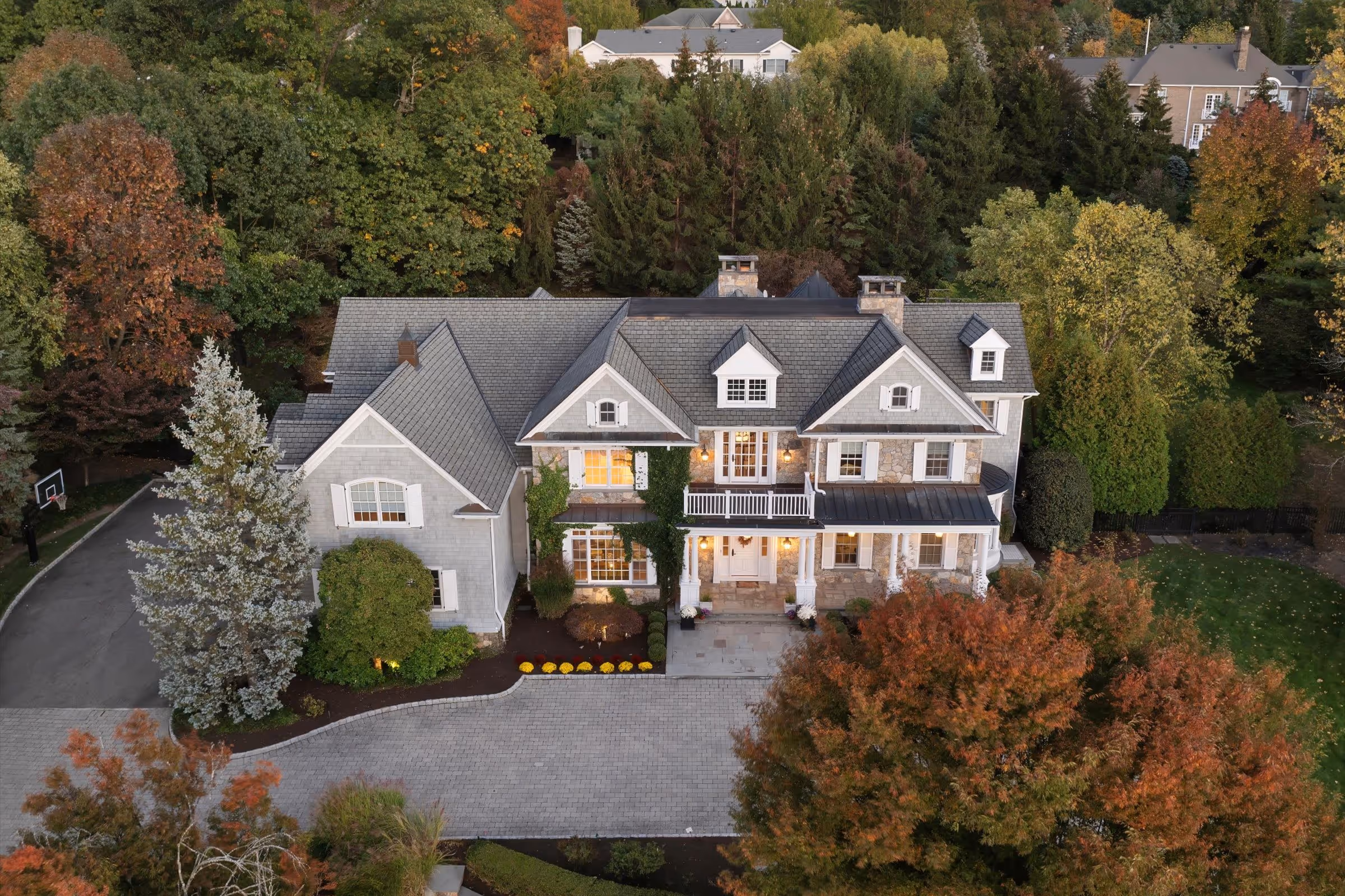 Large gray two-story house with a stone facade, surrounded by green and autumn-colored trees, with a paved driveway and front porch lights on.