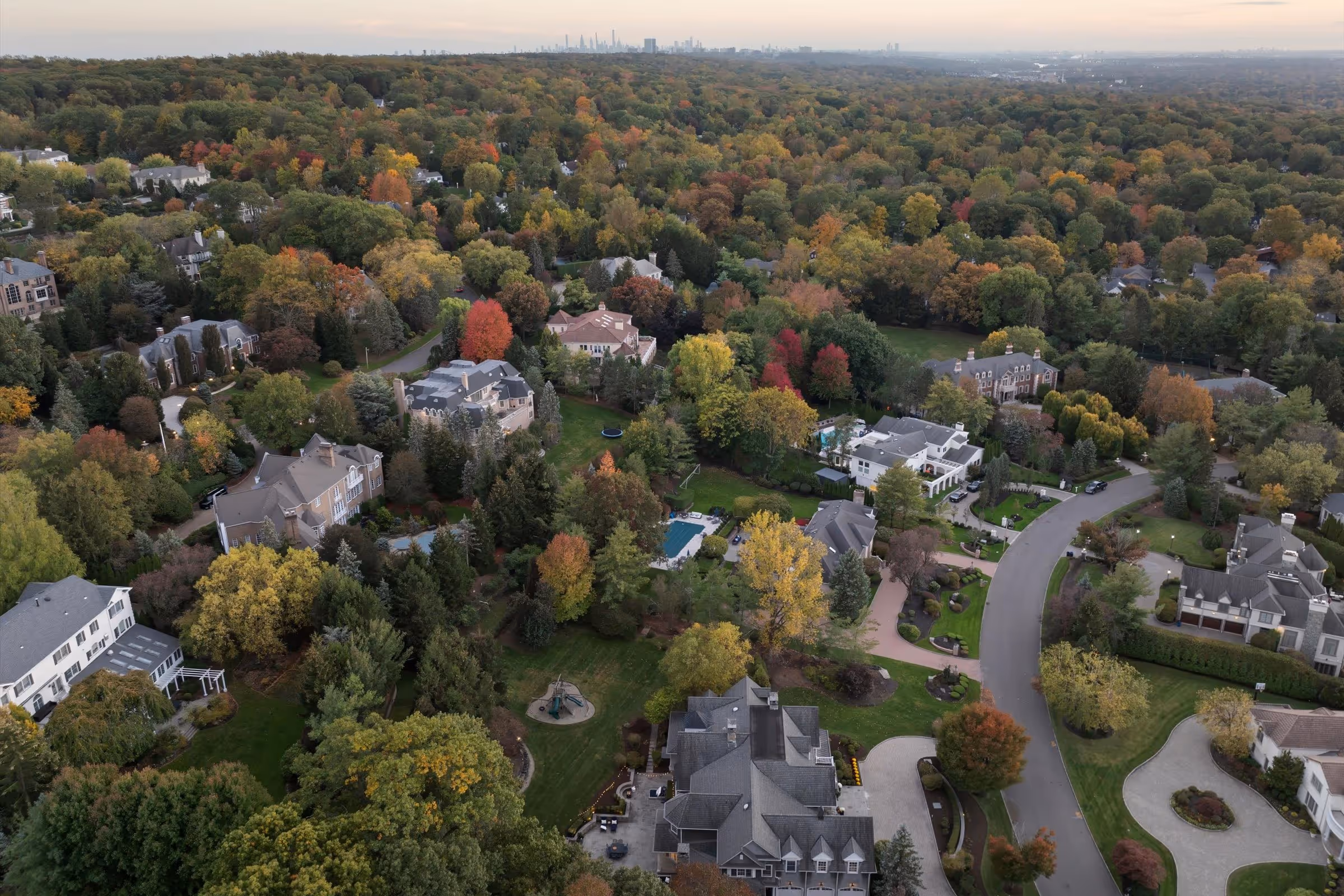 Aerial view of a suburban neighborhood with large houses, colorful autumn trees, green lawns, and winding roads.