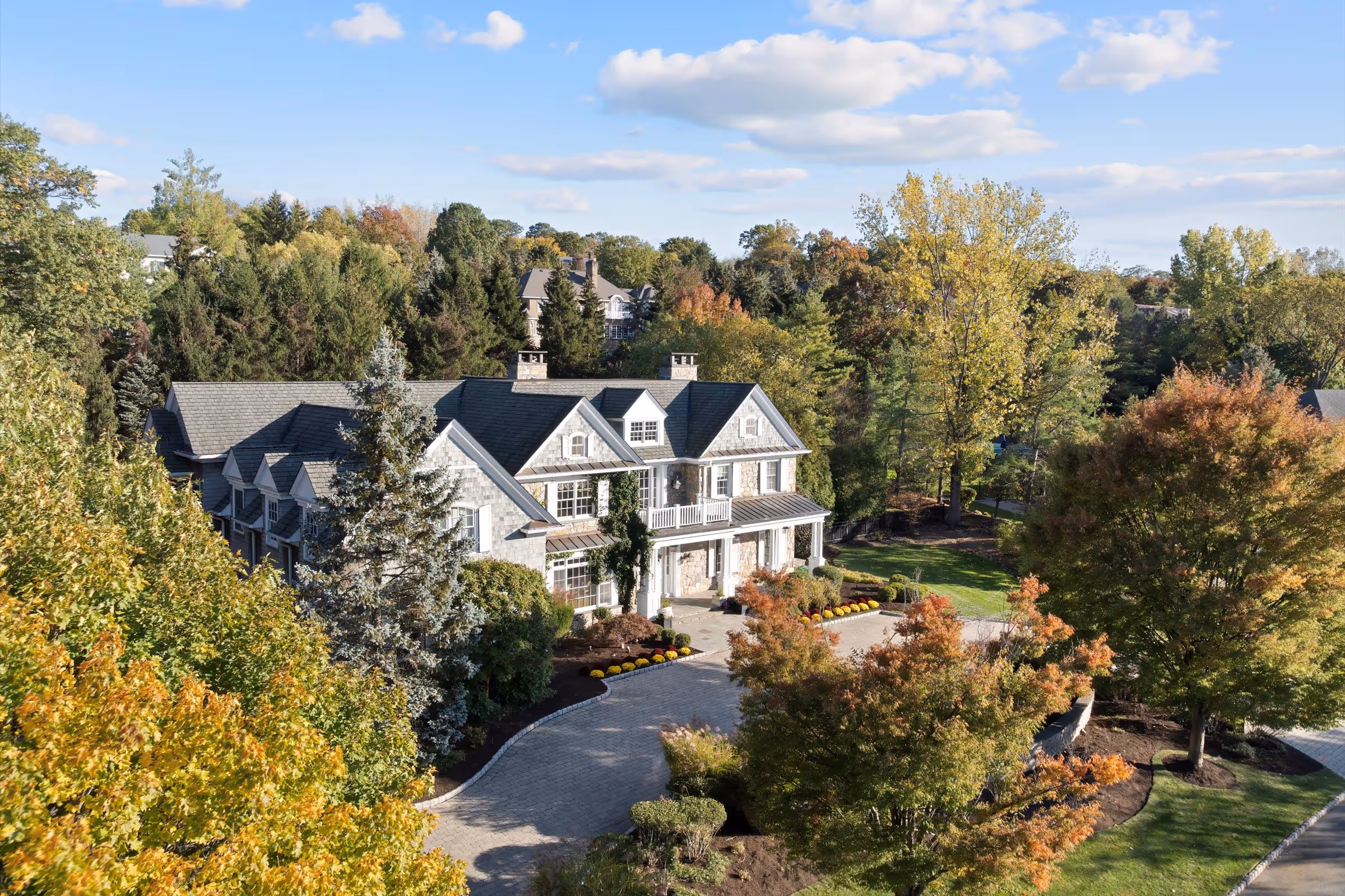 Large two-story house with stone and shingle siding surrounded by trees with autumn foliage under a partly cloudy sky.