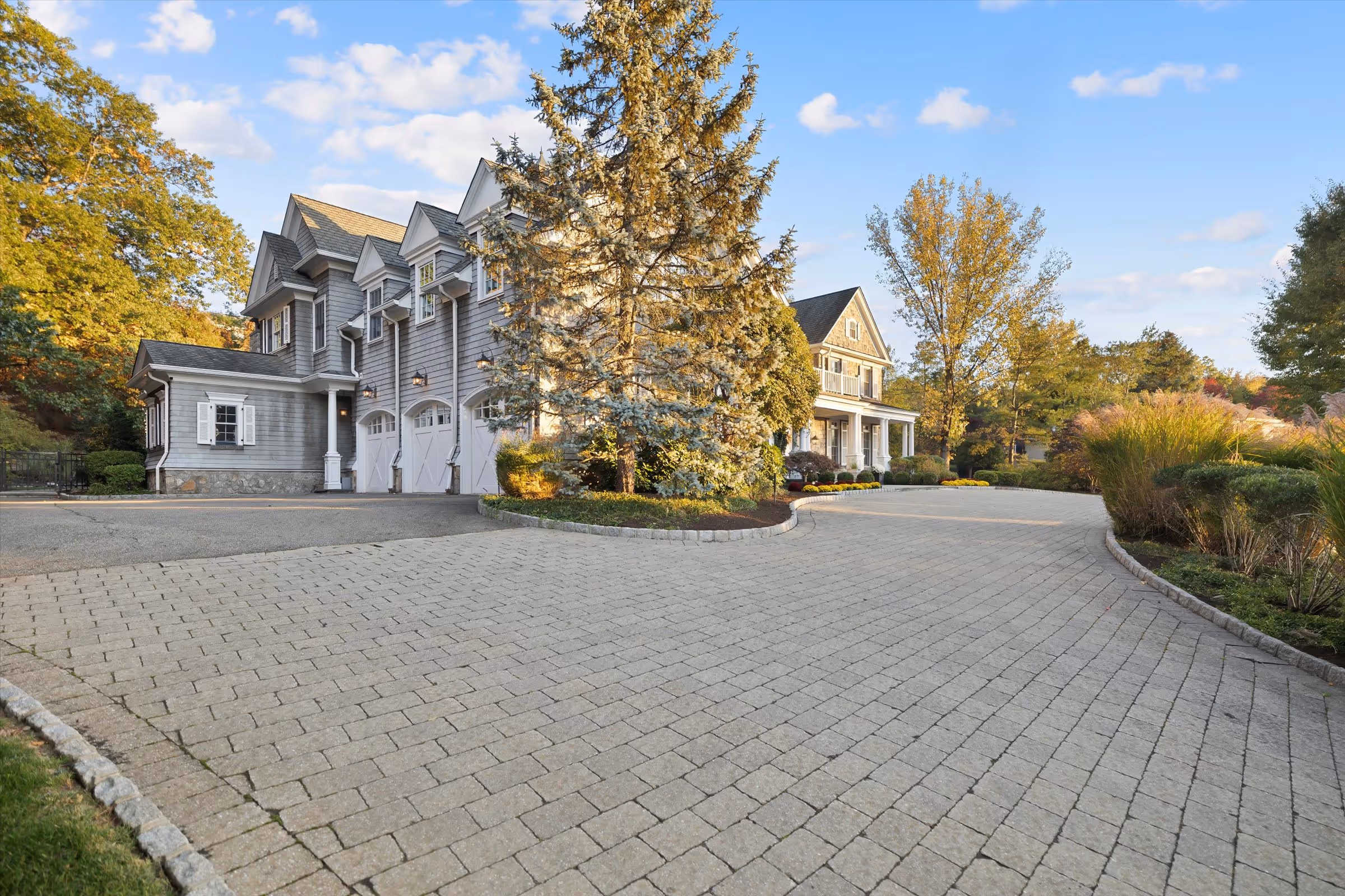 Large gray house with white garage doors, surrounded by trees and a paved driveway under a partly cloudy sky.
