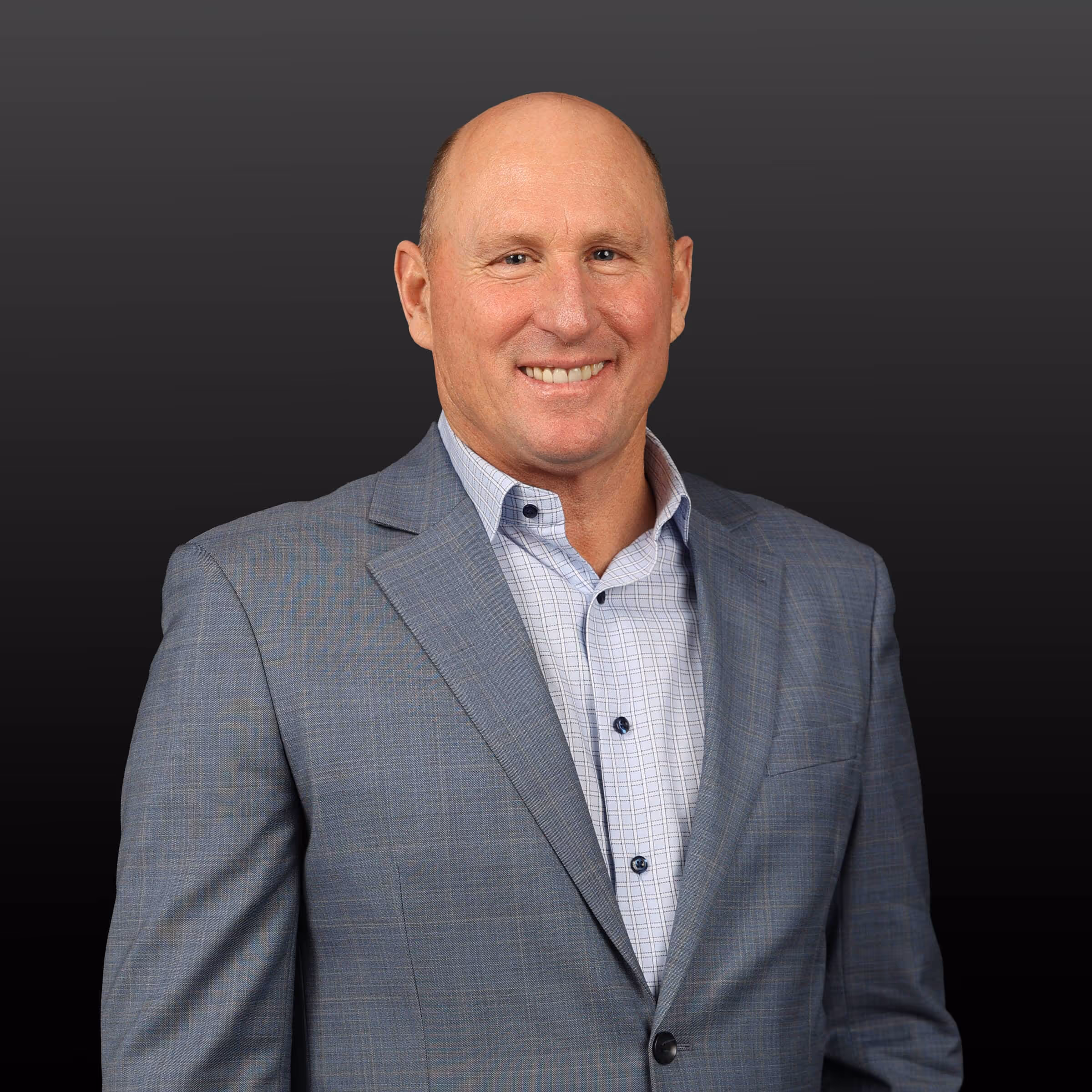 Smiling bald man in a gray plaid suit jacket and white checkered shirt against a dark background.