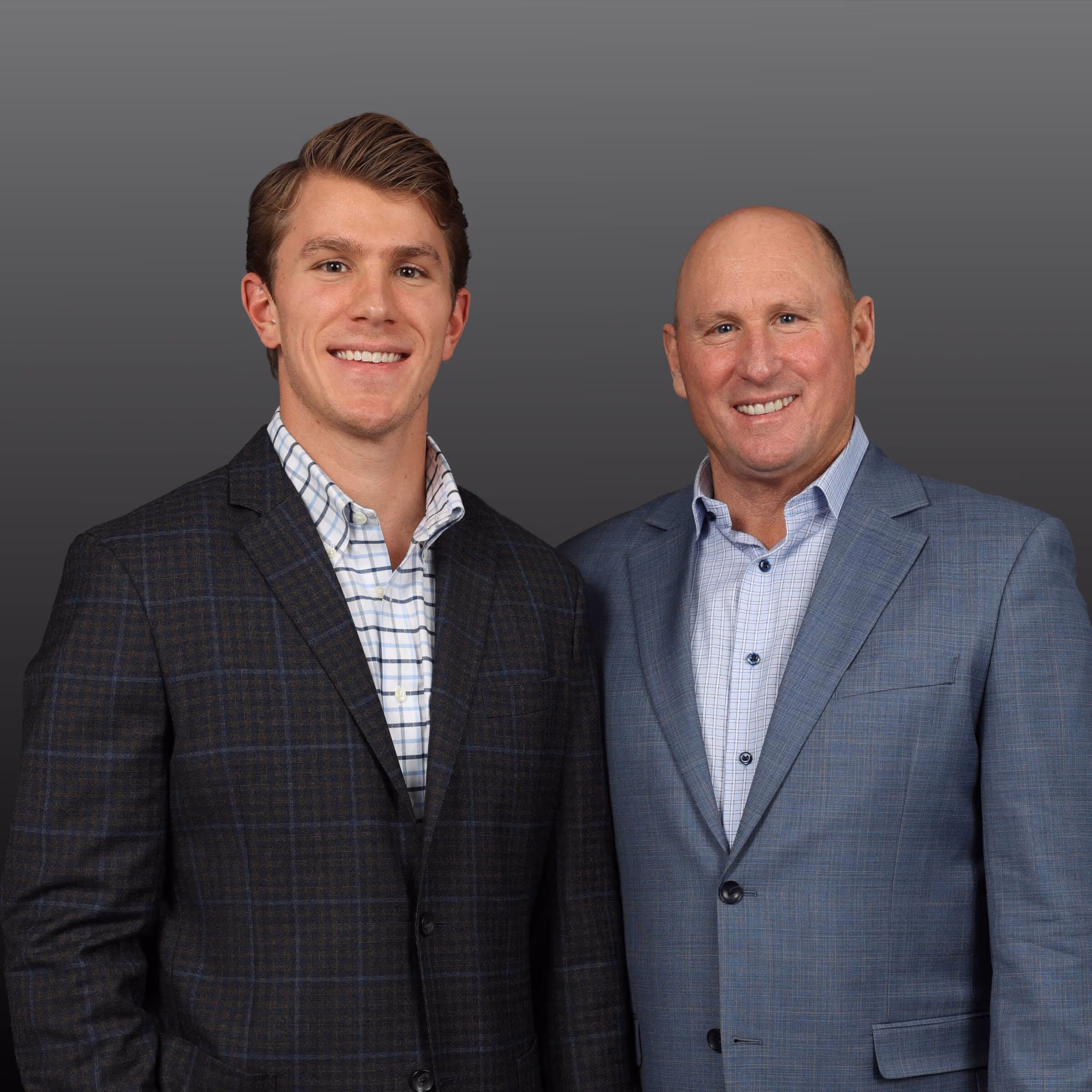 Two men wearing blazers and collared shirts standing side by side smiling against a gray background.