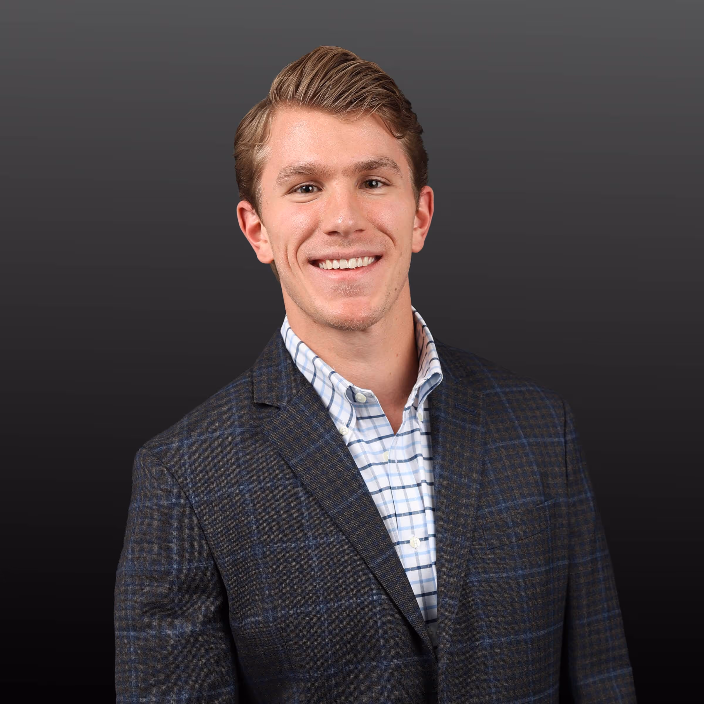 Smiling young man wearing a dark plaid blazer and checkered shirt against a gradient gray background.
