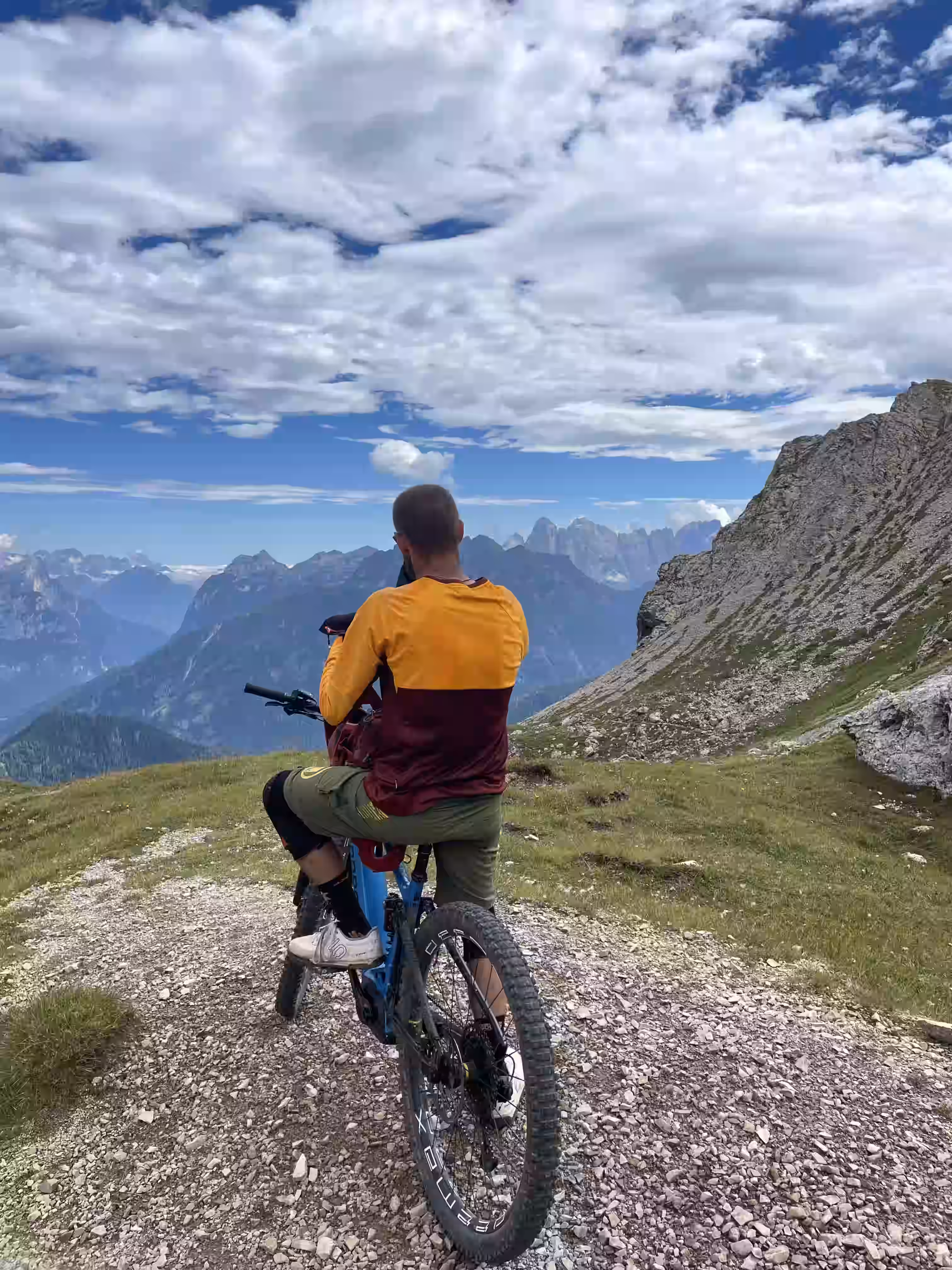 Persona in maglia arancione e marrone su una bicicletta da montagna su un sentiero dolomitico con vista sulle montagne e cielo. Tour guidato nelle dolomiti.