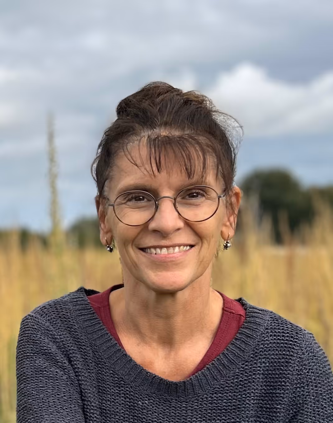 Smiling woman with glasses and bangs wearing a gray sweater over a maroon shirt outdoors with blurred field and cloudy sky background.