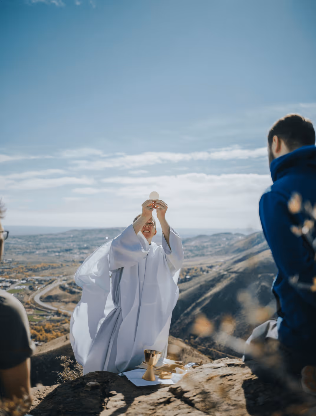 A priest in white robes holds up a communion wafer during an outdoor religious ceremony on a hill overlooking a city.