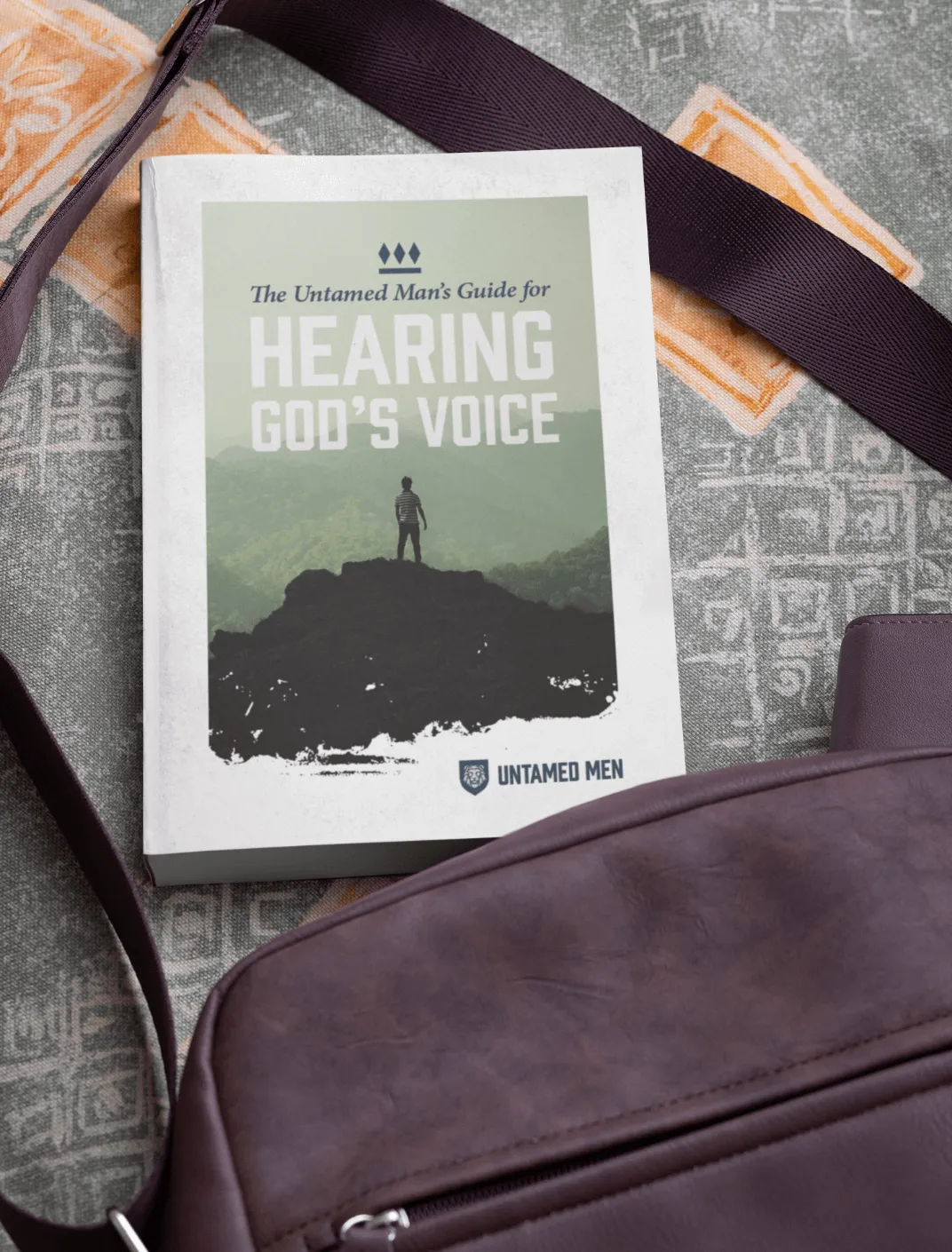 Book titled 'The Untamed Man's Guide for Hearing God's Voice' resting on a patterned surface next to a purple bag.