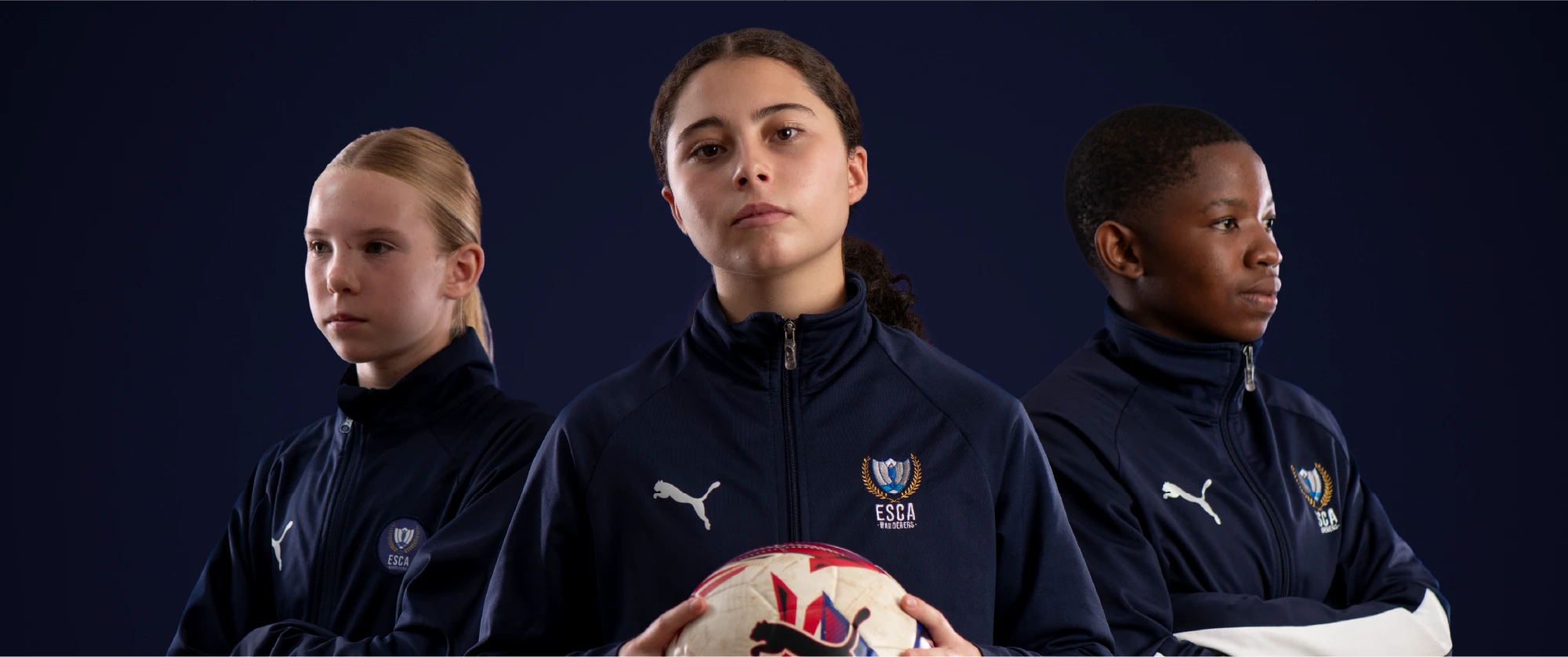 Three young soccer players in navy ESCA jerseys standing confidently against a dark background, center player holding a soccer ball.