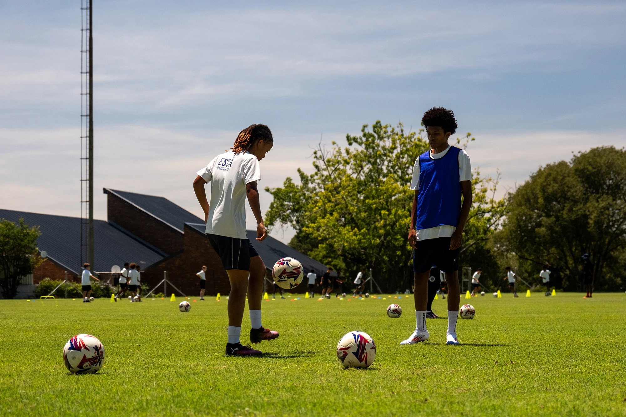 Two young soccer players practicing on a green field with several soccer balls and cones, under a partly cloudy sky.