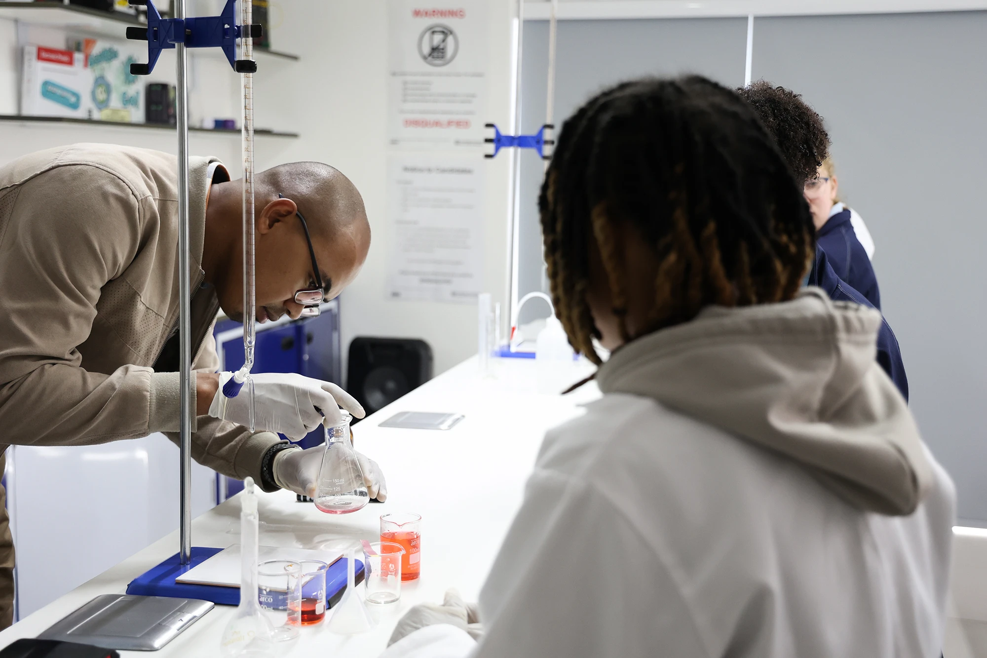 ESCA Scientist wearing gloves carefully pouring liquid from a pipette into a flask in a laboratory with students observing.