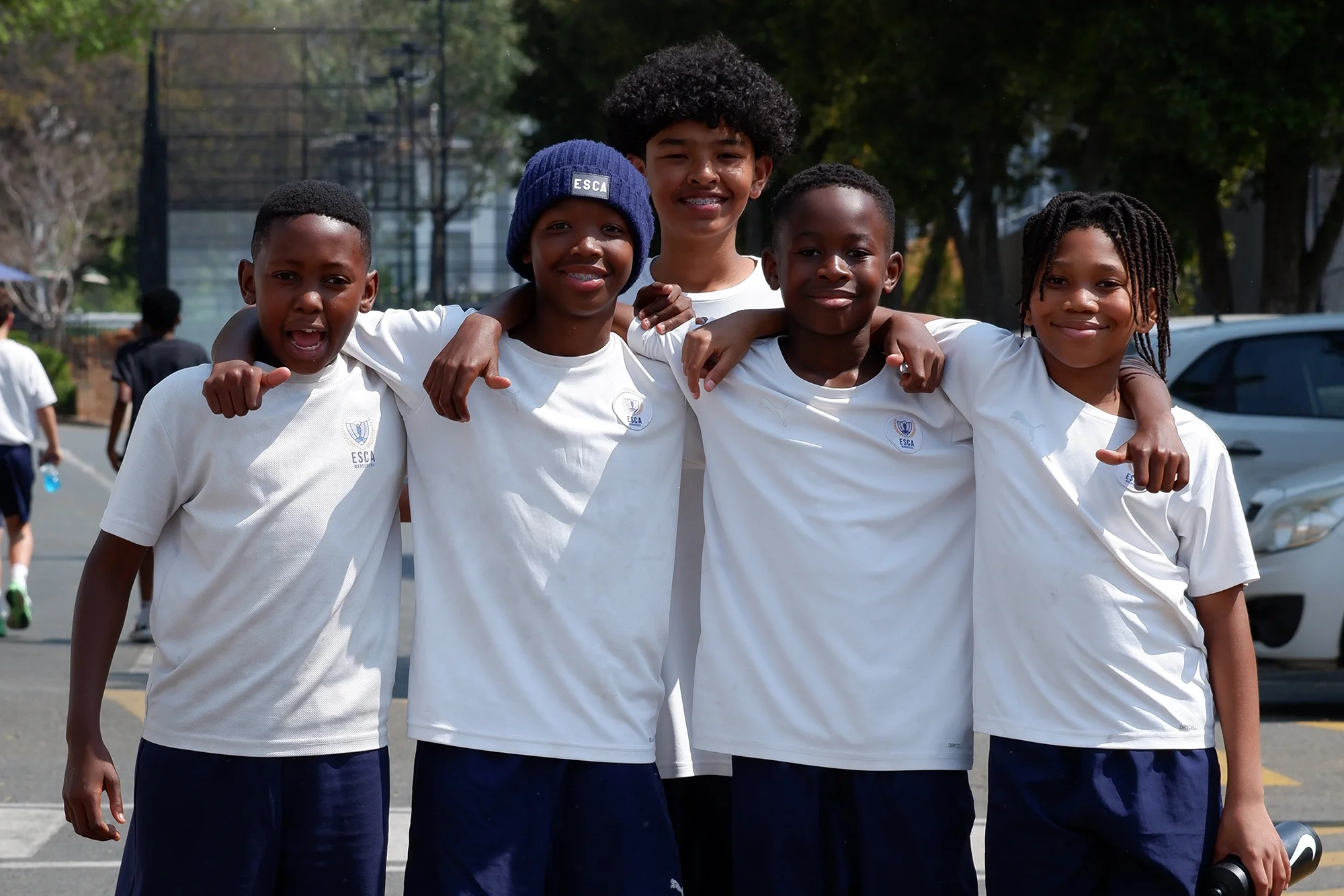 Five boys in white t-shirts and navy shorts standing closely with arms around each other, smiling outdoors on a sunny day.