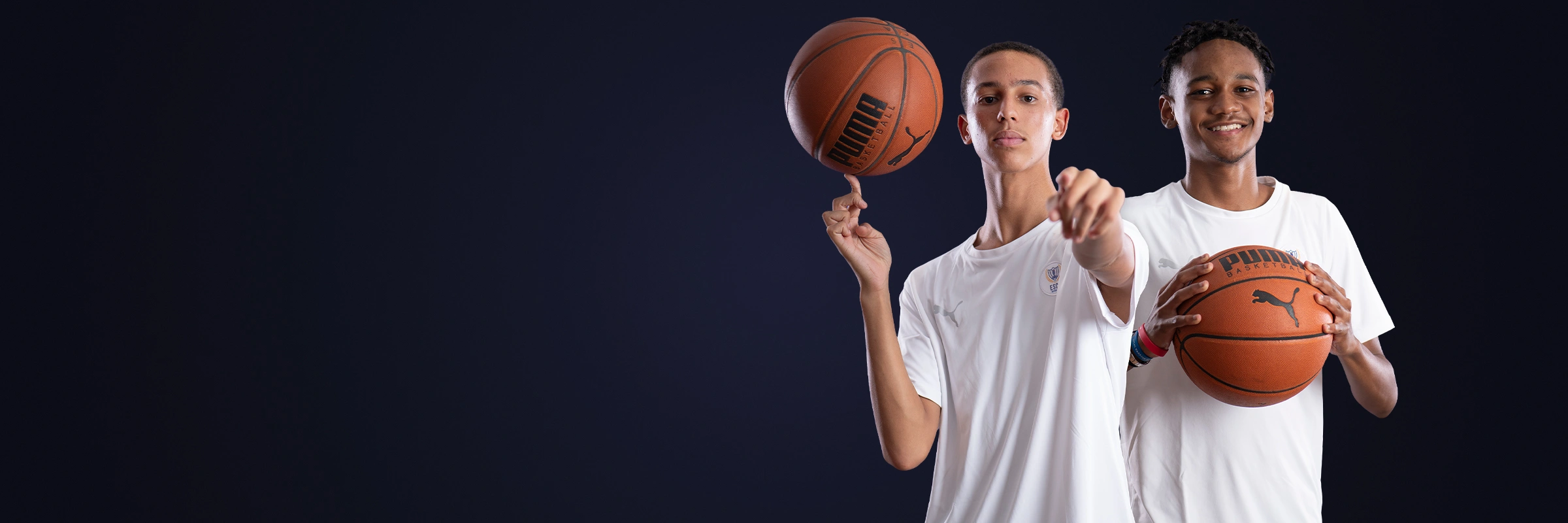 Two young men in white shirts holding basketballs against a dark background, one spinning a ball on his finger and the other smiling while holding a basketball.