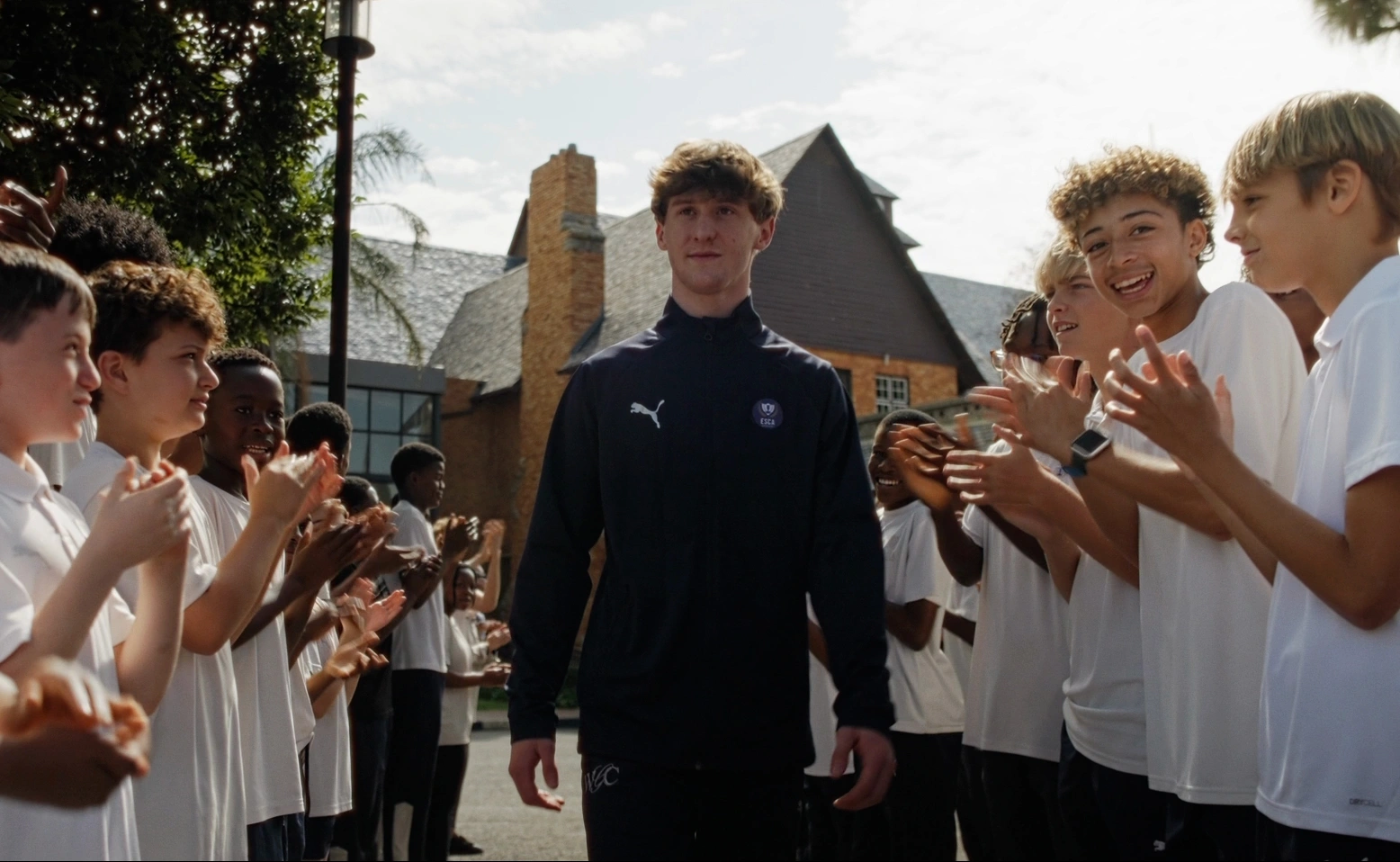 Five boys in white t-shirts and navy shorts standing closely with arms around each other, smiling outdoors on a sunny day.