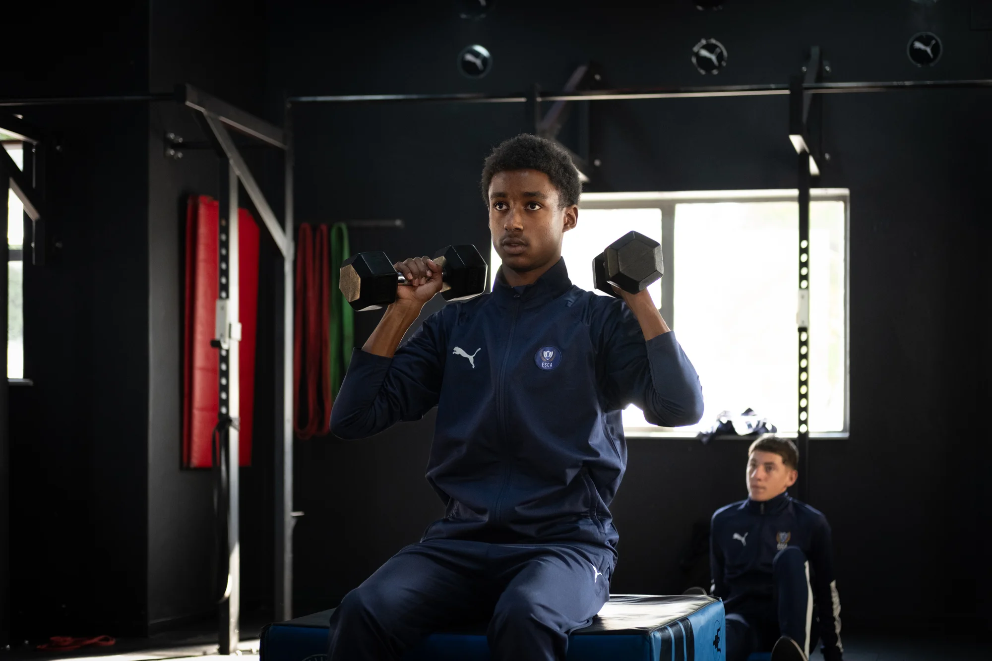 Five boys in white t-shirts and navy shorts standing closely with arms around each other, smiling outdoors on a sunny day.