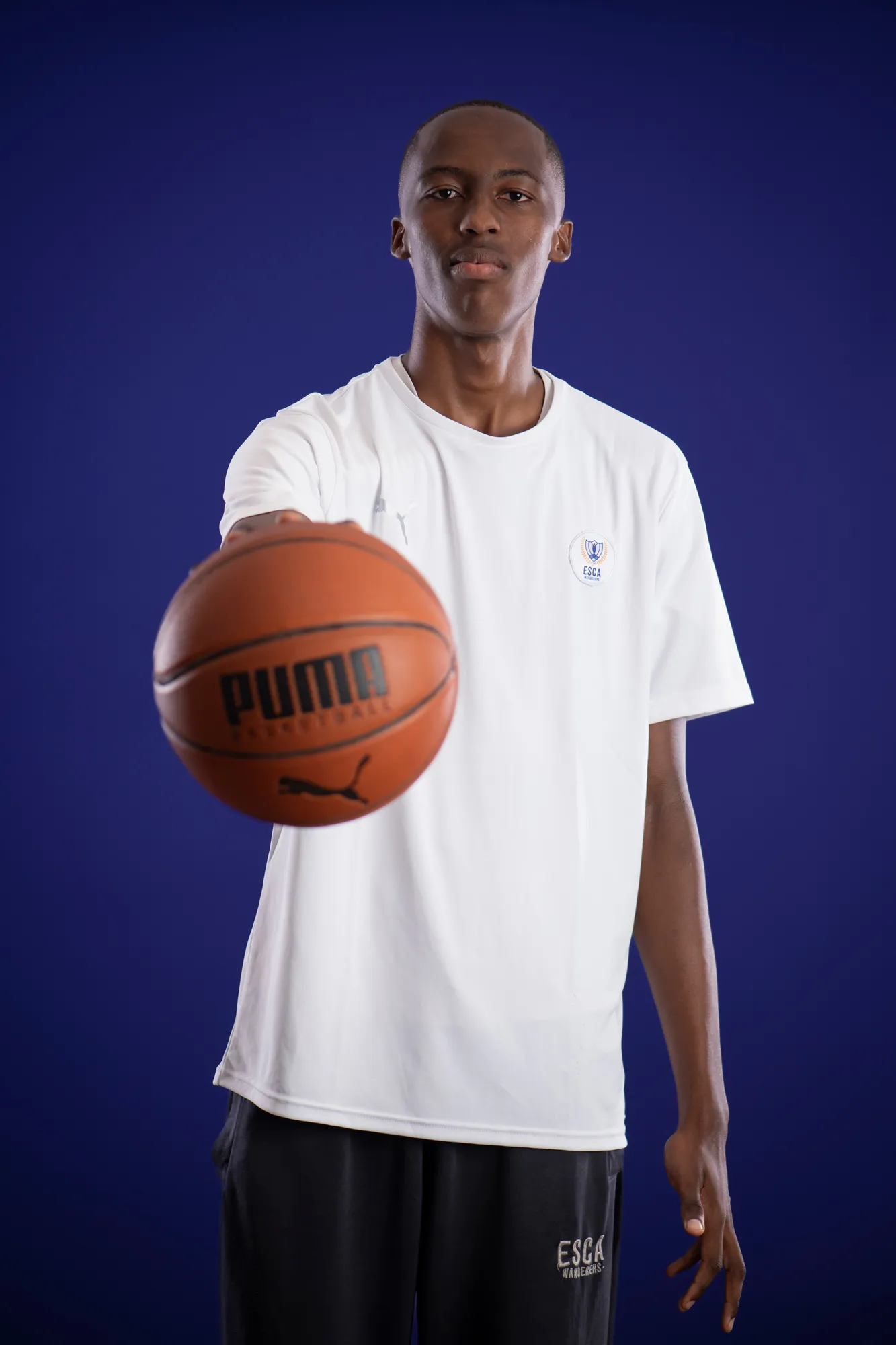 A young man in a white ESCA t-shirt holding a basketball towards the camera against a dark blue background.